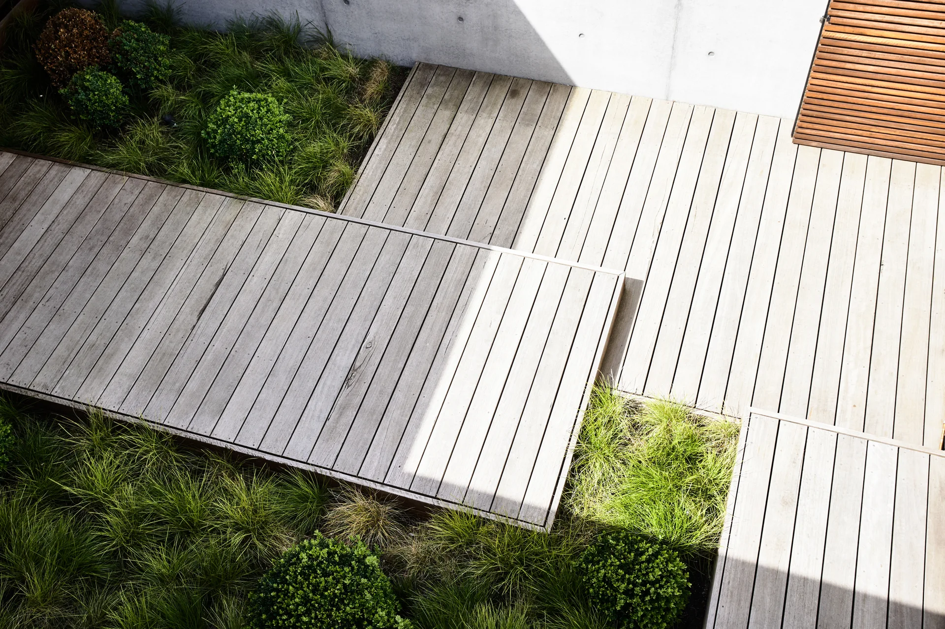 An overhead view of tiered, light brown wooden decking platforms surrounded by green ornamental grass and a smooth grey concrete retaining wall.