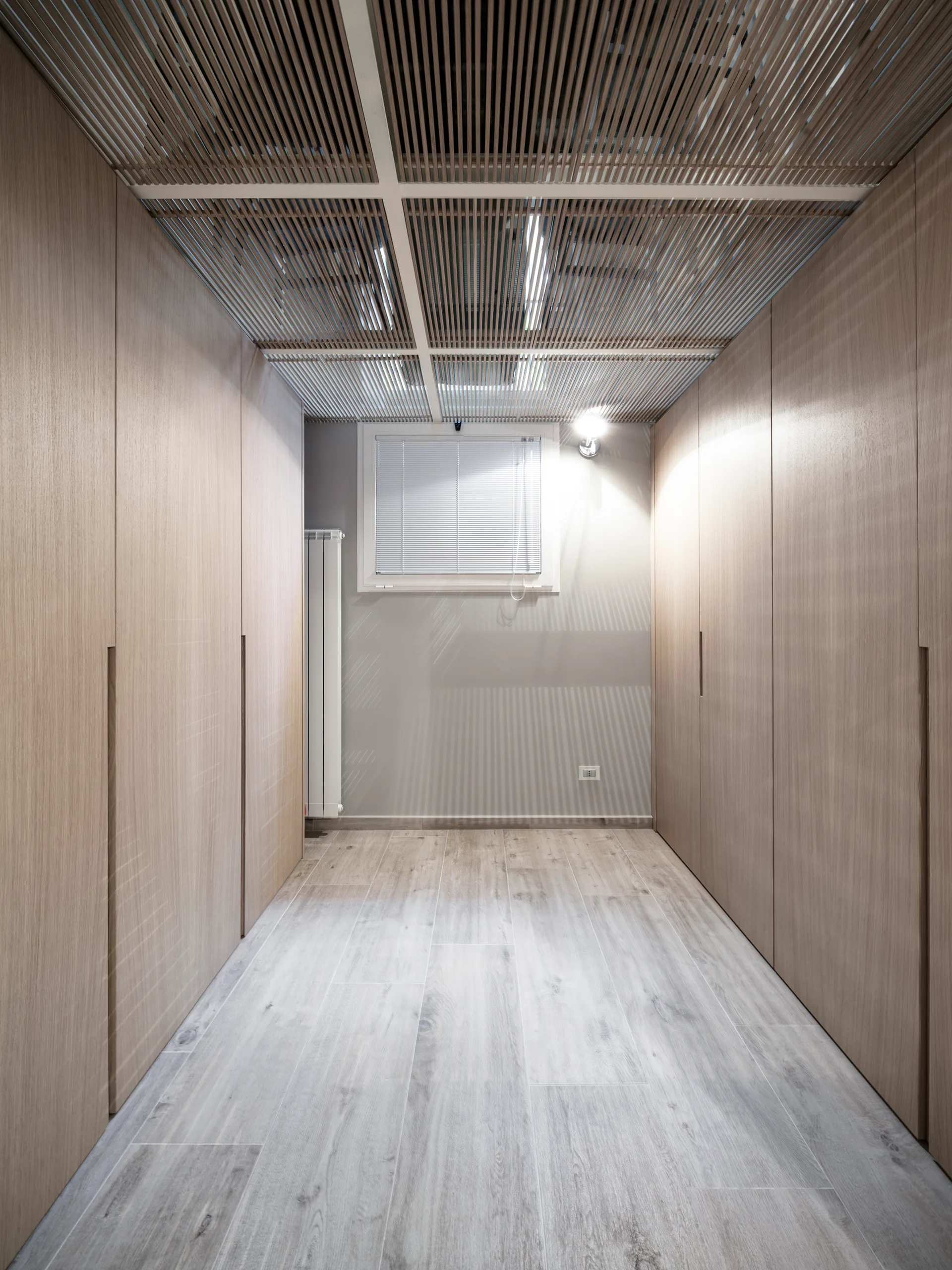 A symmetrical dressing room with floor-to-ceiling wooden closets on both sides, a slatted timber ceiling, and a small back window with blinds.