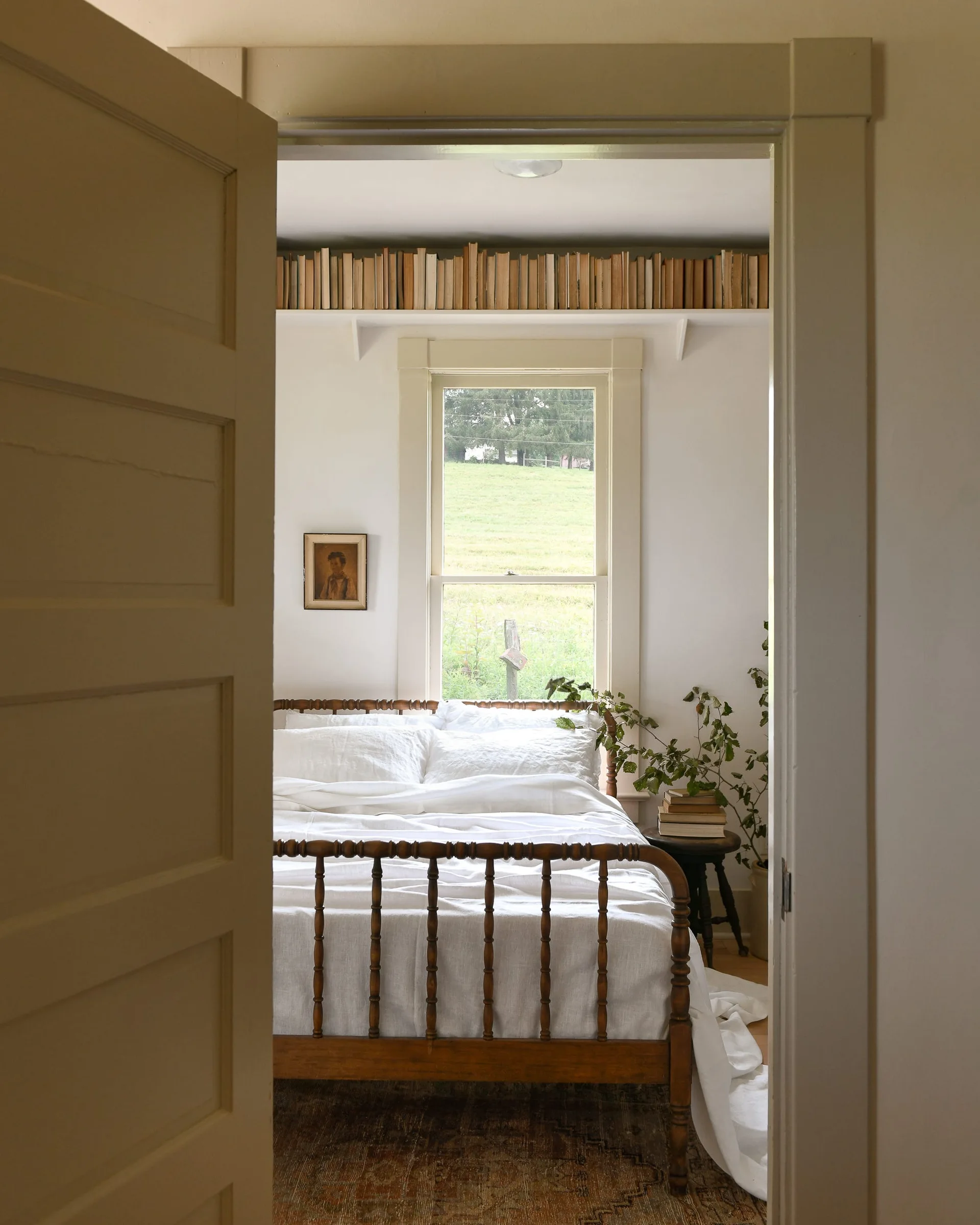 A view through an open doorway into a farmhouse bedroom featuring a wooden spindle bed and a high shelf filled with books above the window.