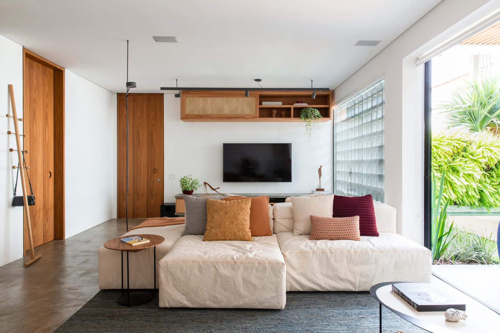 A bright living room features a cream sectional sofa, a wall-mounted TV under a wooden cane cabinet, and a large glass block window.