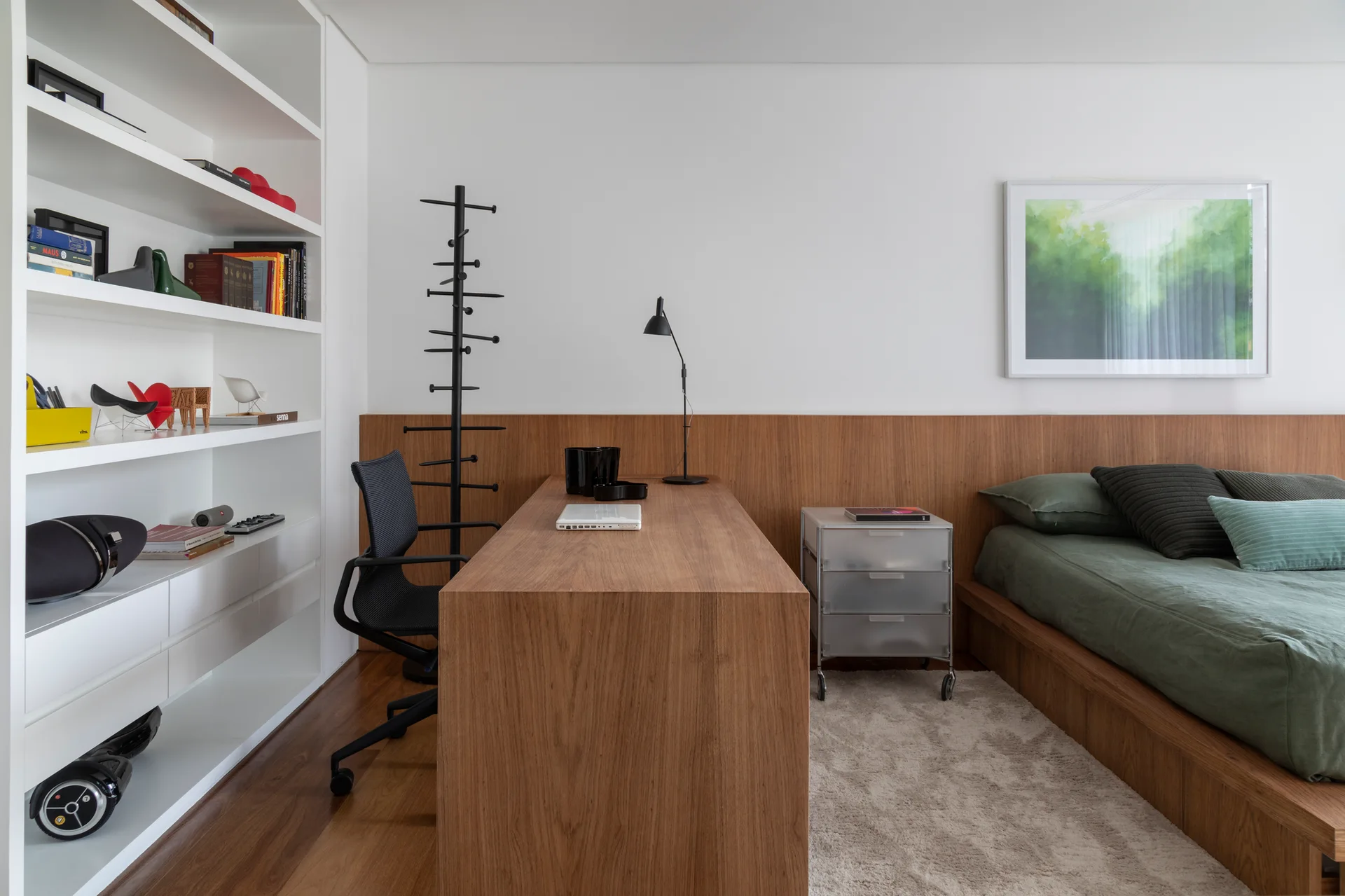 A modern bedroom features a low wooden platform bed with green bedding, a matching built-in desk, and white shelving.