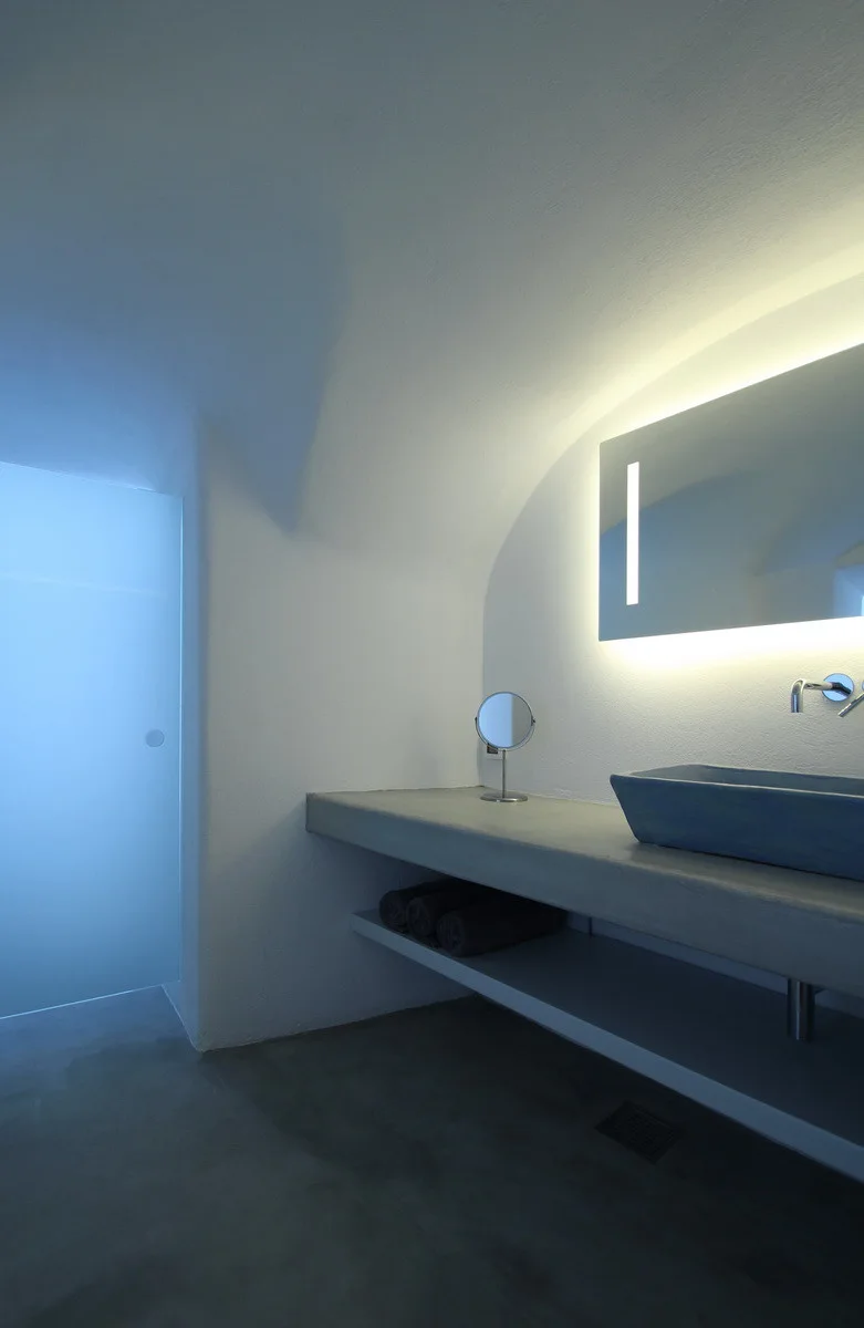 A minimalist bathroom features a floating grey concrete vanity with a blue sink, a backlit mirror, and white curved plaster walls.