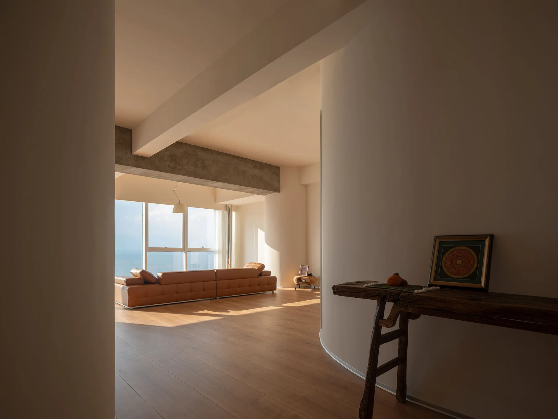 A spacious minimalist room featuring a curved wall, a rustic wood console, and a tan leather sofa facing large windows with a sea view.