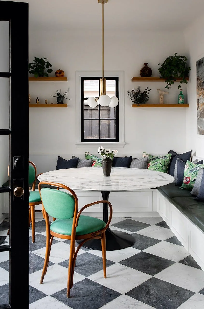 A dining nook features a round marble table, built-in banquette, green bentwood chairs, and a black and white checkered tile floor.