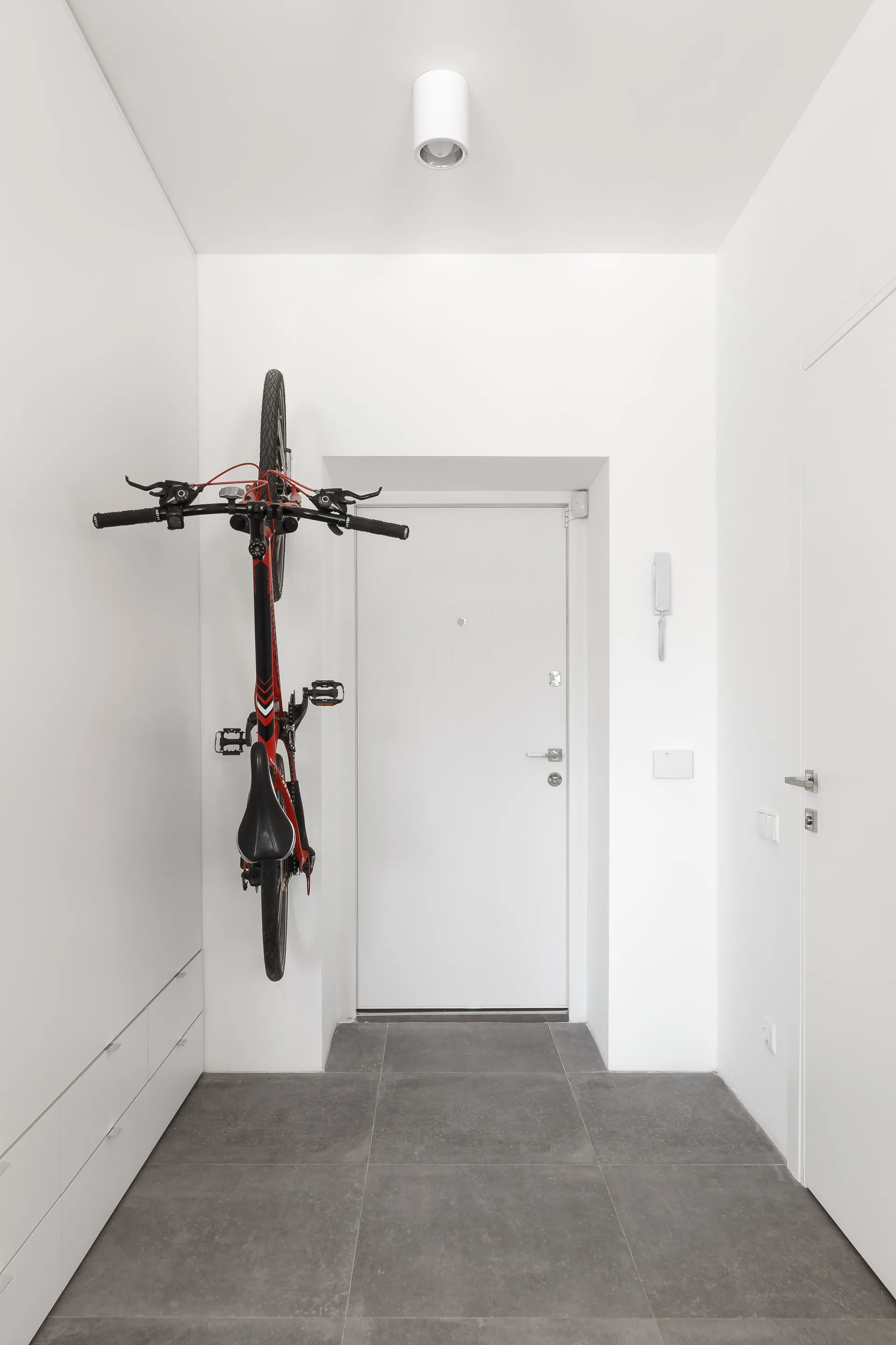 A minimalist white entryway featuring a black and red bicycle mounted vertically on the wall beside grey floor tiles and built-in cabinets.