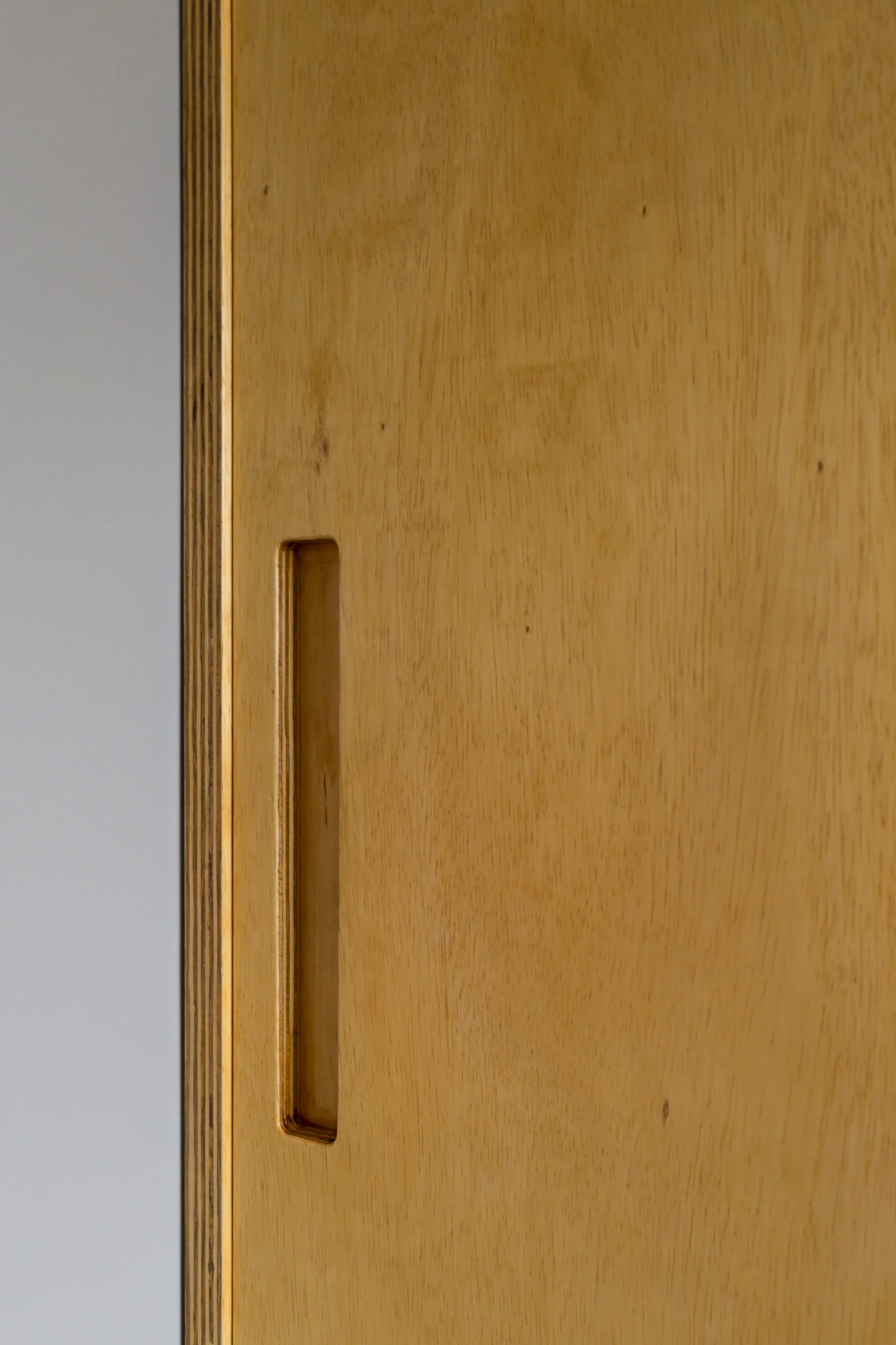 A close-up of a light wood plywood door with an integrated vertical recessed handle and a layered edge detail.