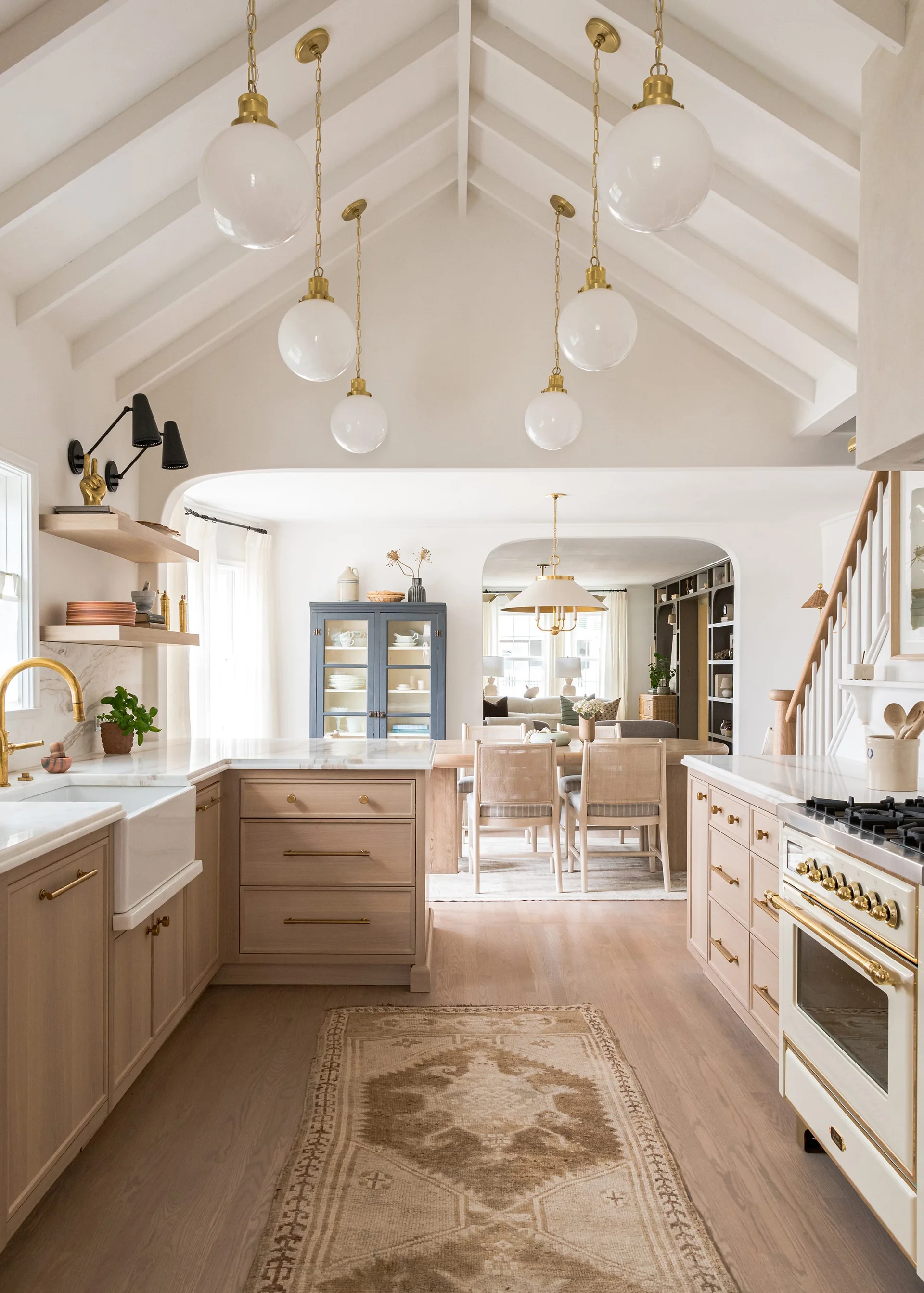 A light wood kitchen with a vaulted beam ceiling, globe pendant lights, and a farmhouse sink, opening into a dining room through an archway.