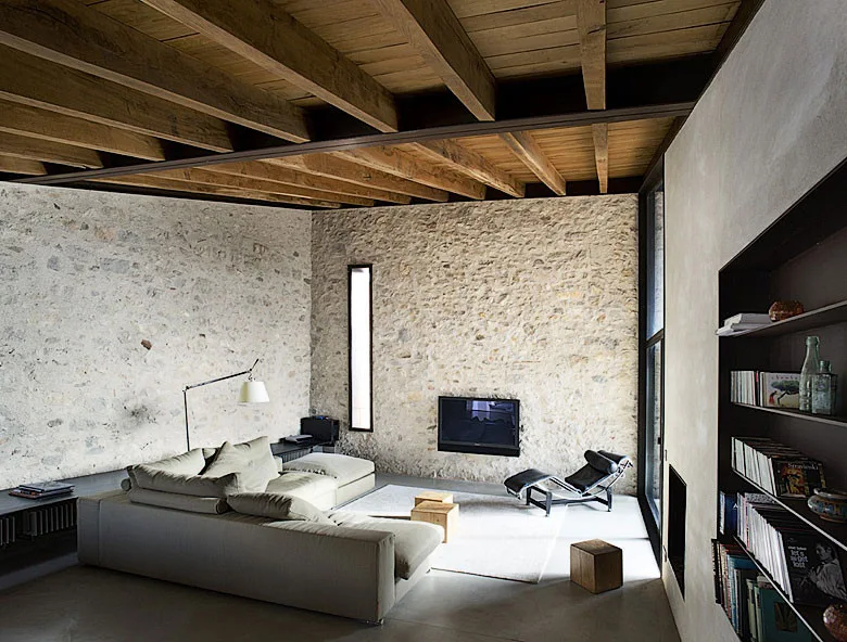 A spacious living room with natural stone walls, a wood plank ceiling with beams, a beige sectional sofa, and a black leather lounge chair.