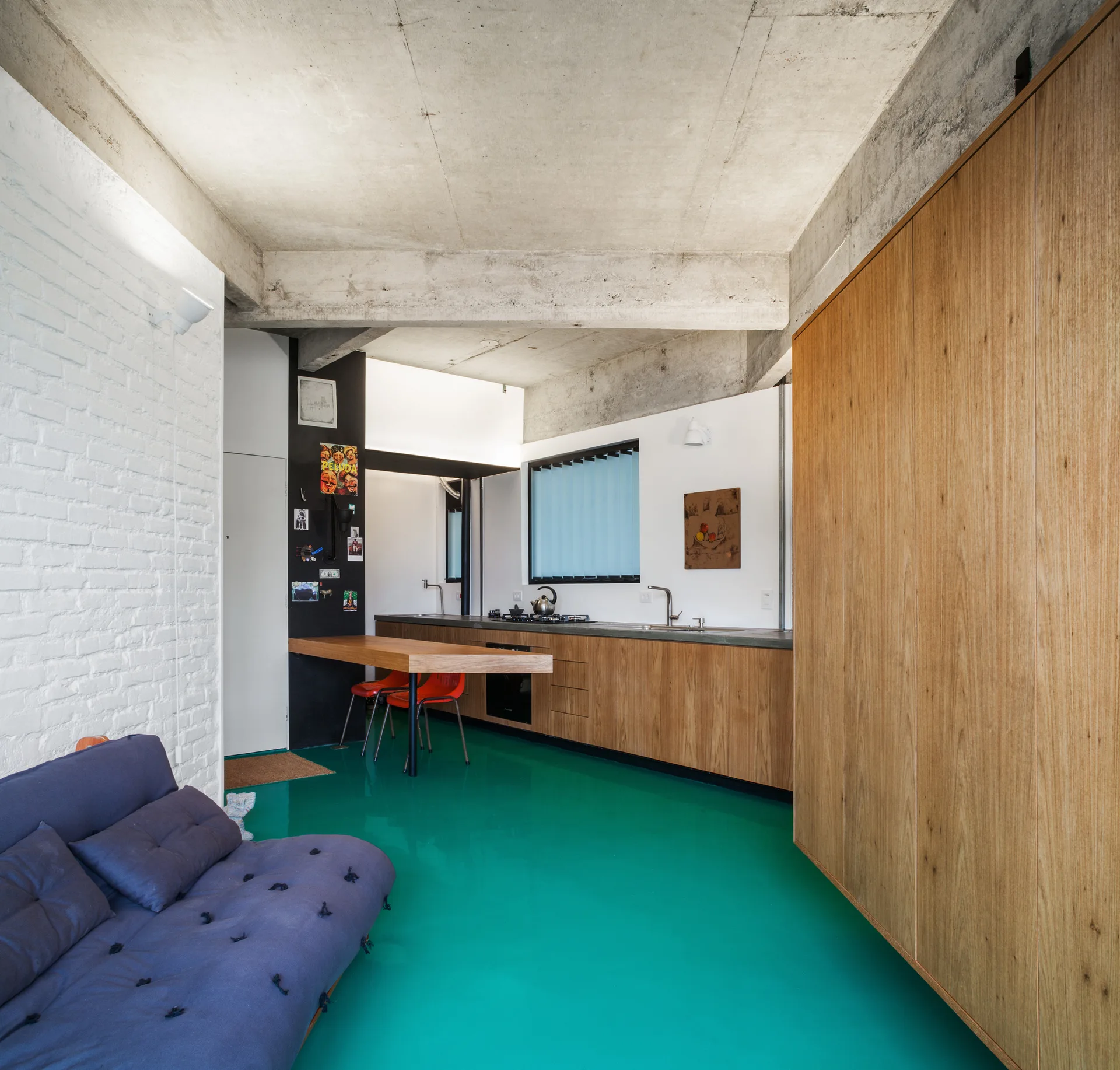 A modern open-plan kitchen features a glossy teal floor, warm wood cabinets, a white brick wall, and a raw concrete ceiling with exposed beams.