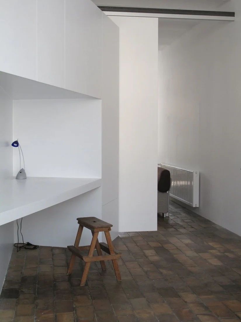 A minimalist home office features a white built-in desk, a small wooden step stool, and rustic dark brick flooring.