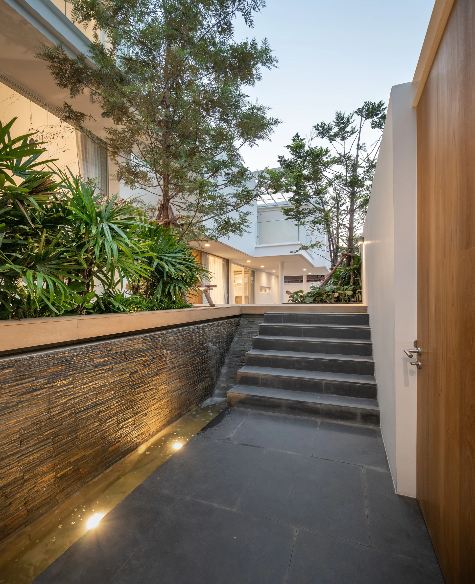 A modern outdoor courtyard featuring a dark stone staircase alongside an illuminated stacked-stone water feature and lush green planters.