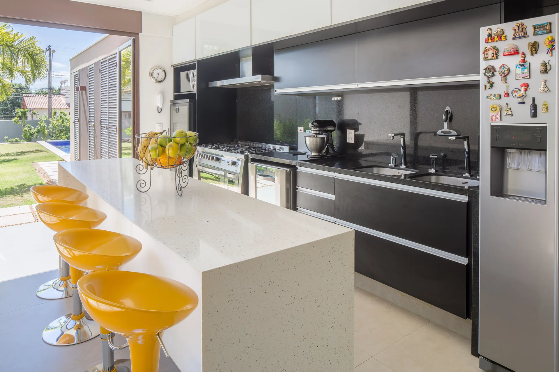 A modern kitchen features a long white quartz island lined with four glossy yellow bar stools, set against black cabinets and a sliding glass door.