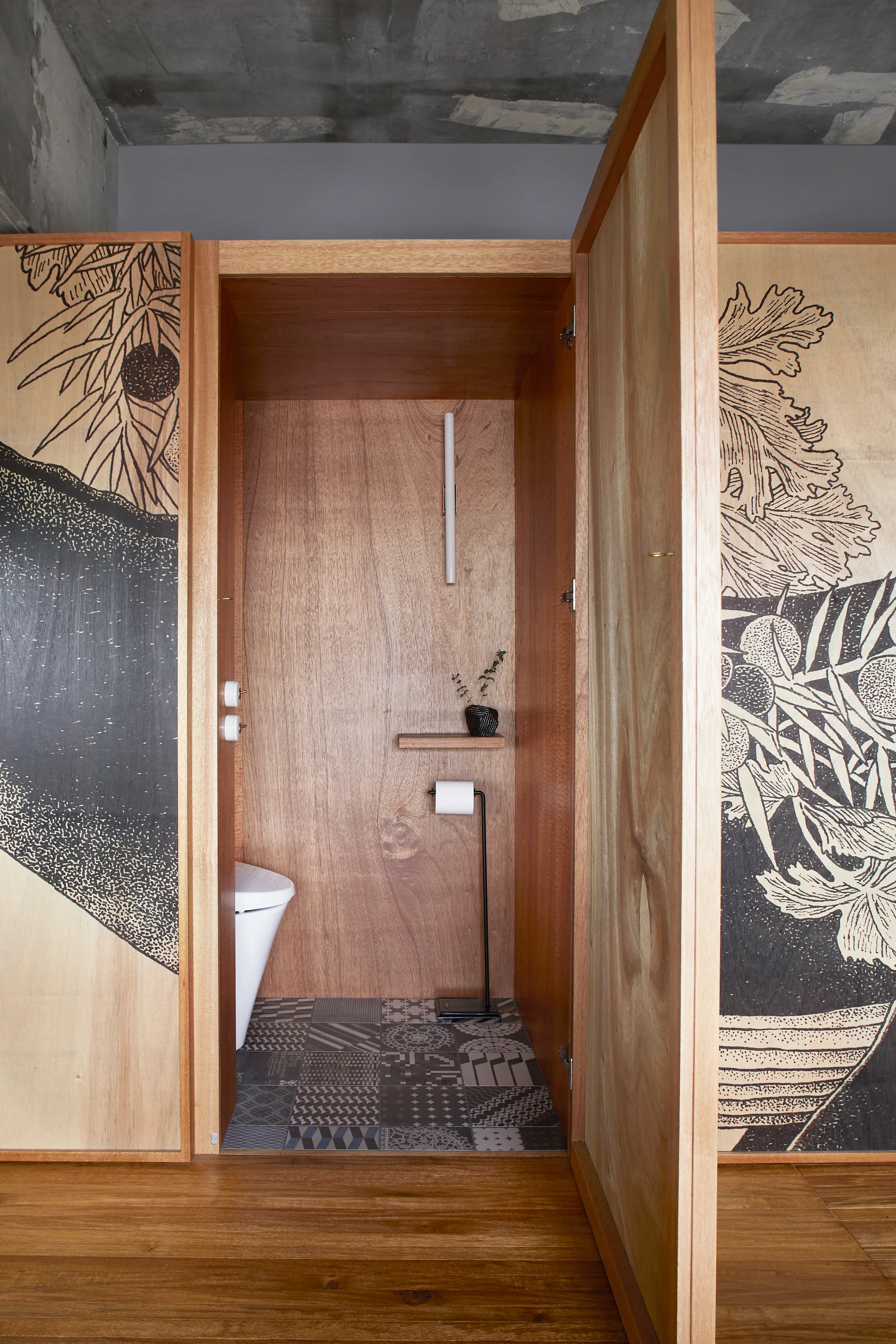 A compact powder room featuring warm wood paneled walls, patterned floor tiles, an exposed concrete ceiling, and custom illustrated doors.