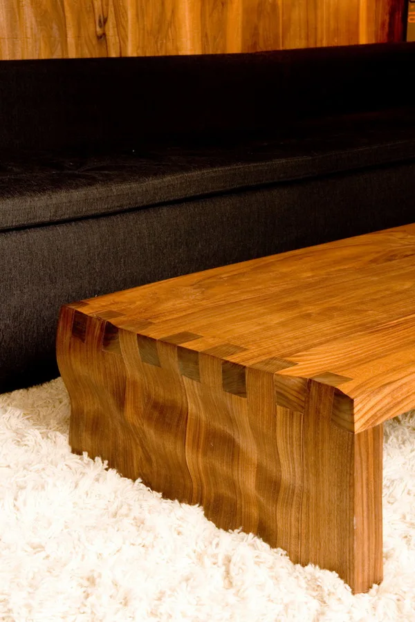 A close-up of a natural wood coffee table with box joints resting on a white shag rug in front of a dark upholstered sofa.