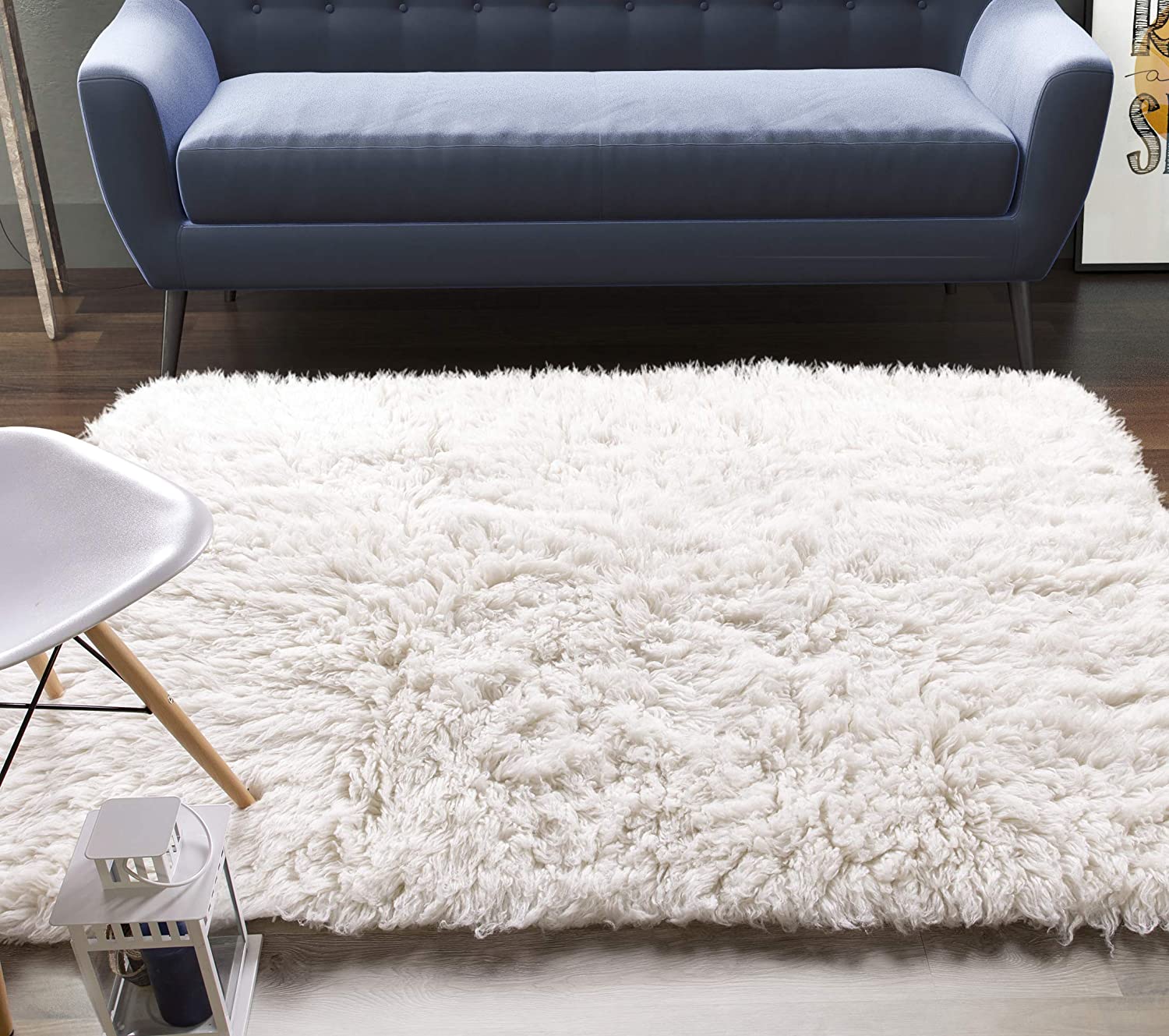 A blue mid-century modern sofa sits behind a thick white shaggy rug on dark wood floors next to a white molded plastic chair.