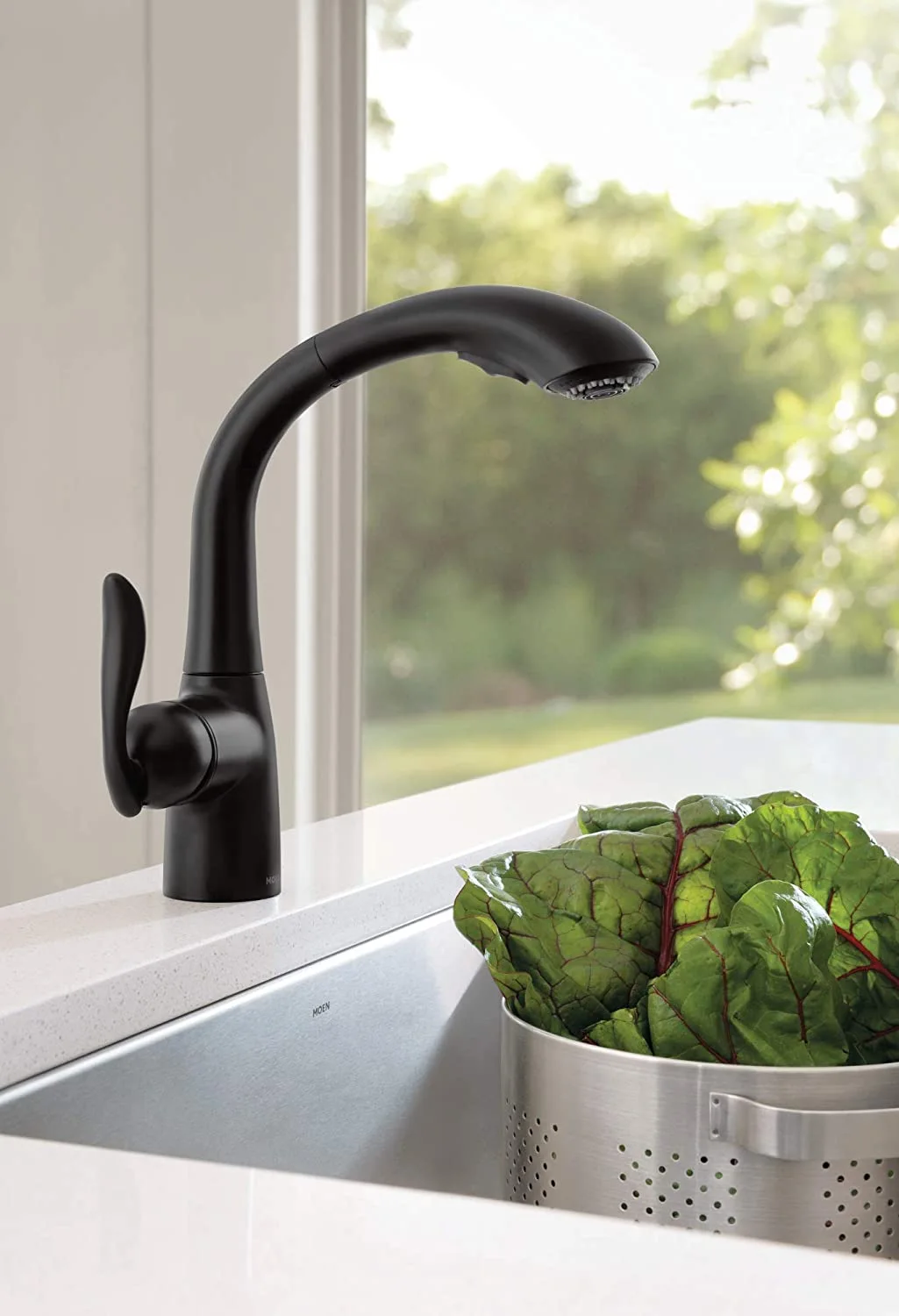 A matte black pull-down kitchen faucet is mounted on a white stone countertop next to a stainless steel sink containing a metal colander of green chard.