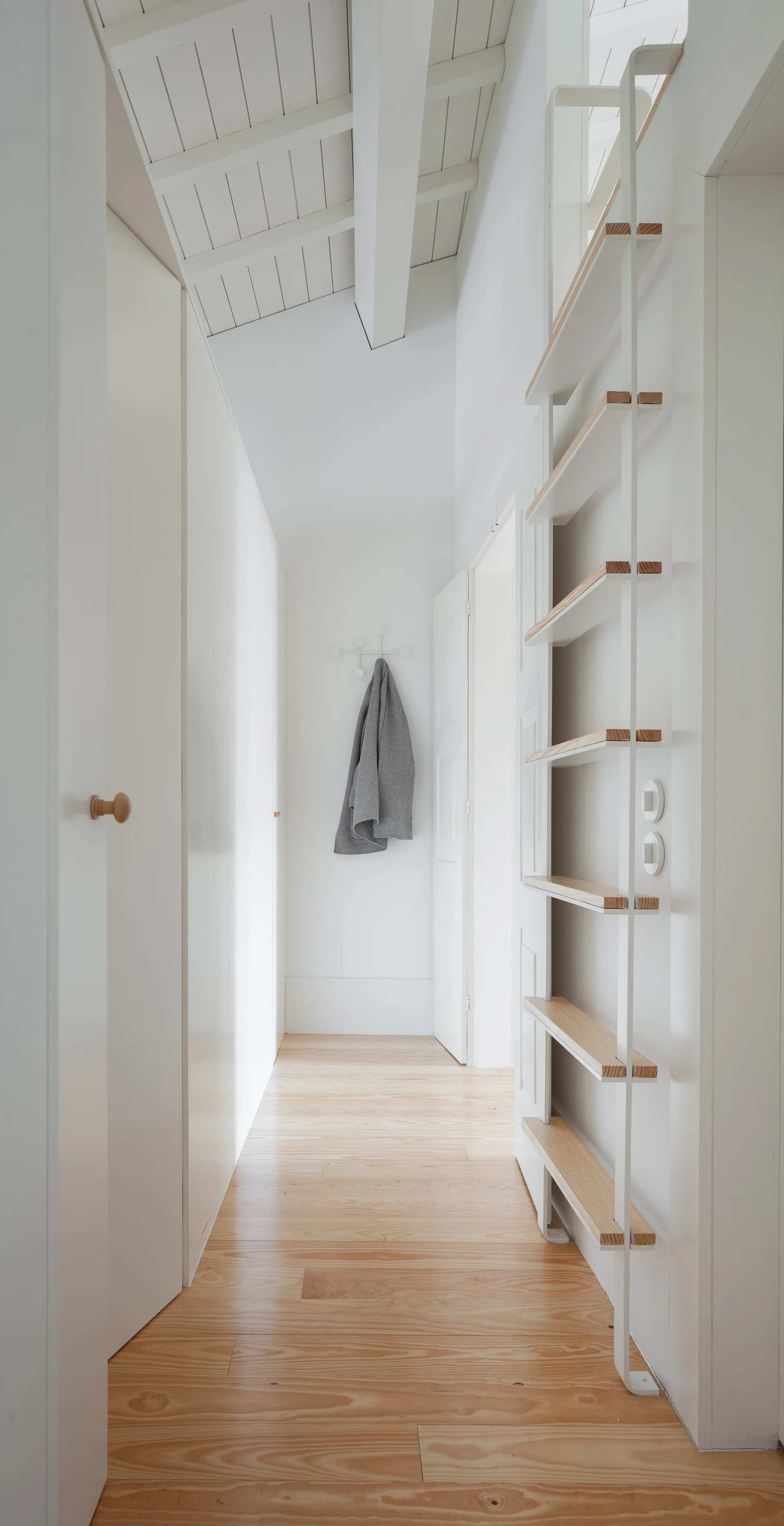 A narrow white hallway featuring light wood flooring, a vaulted ceiling with white beams, and a slim ladder shelf on the wall.