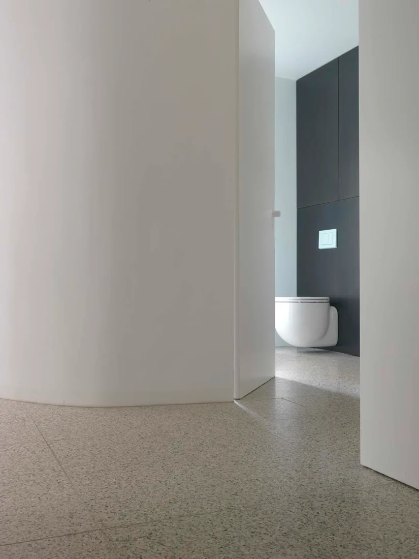 A minimalist bathroom featuring a curved white wall, speckled grey terrazzo flooring, and a white wall-mounted toilet against dark grey paneling.