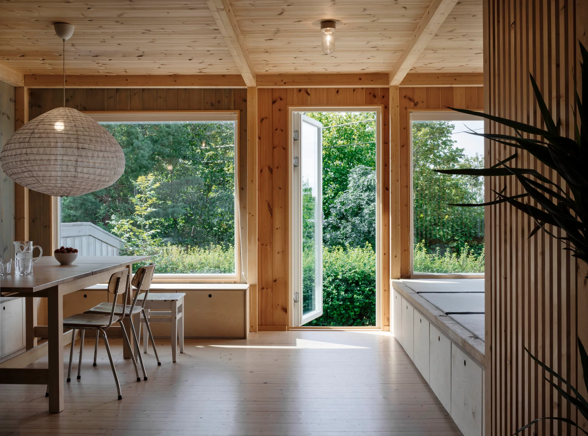 A light wood dining room featuring a wooden table, built-in storage benches, large windows, and an open door looking out to a green garden.