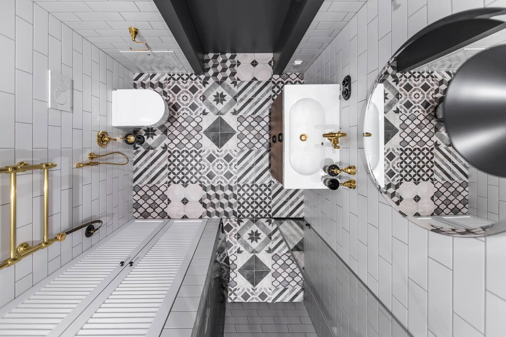 An overhead view of a small bathroom with geometric patterned floor tiles, white subway tile walls, and a wall-mounted toilet with gold hardware.