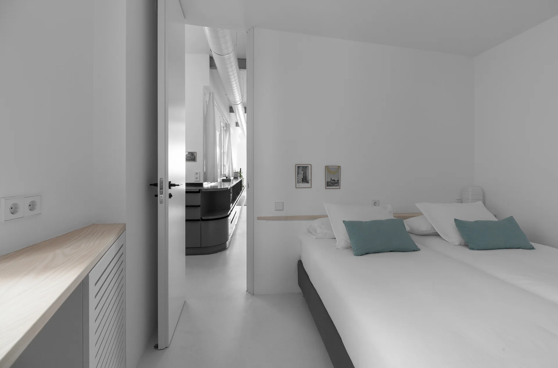 A minimalist white bedroom with twin beds featuring teal pillows, a narrow wood ledge behind the bed, and an open doorway leading to a hall.