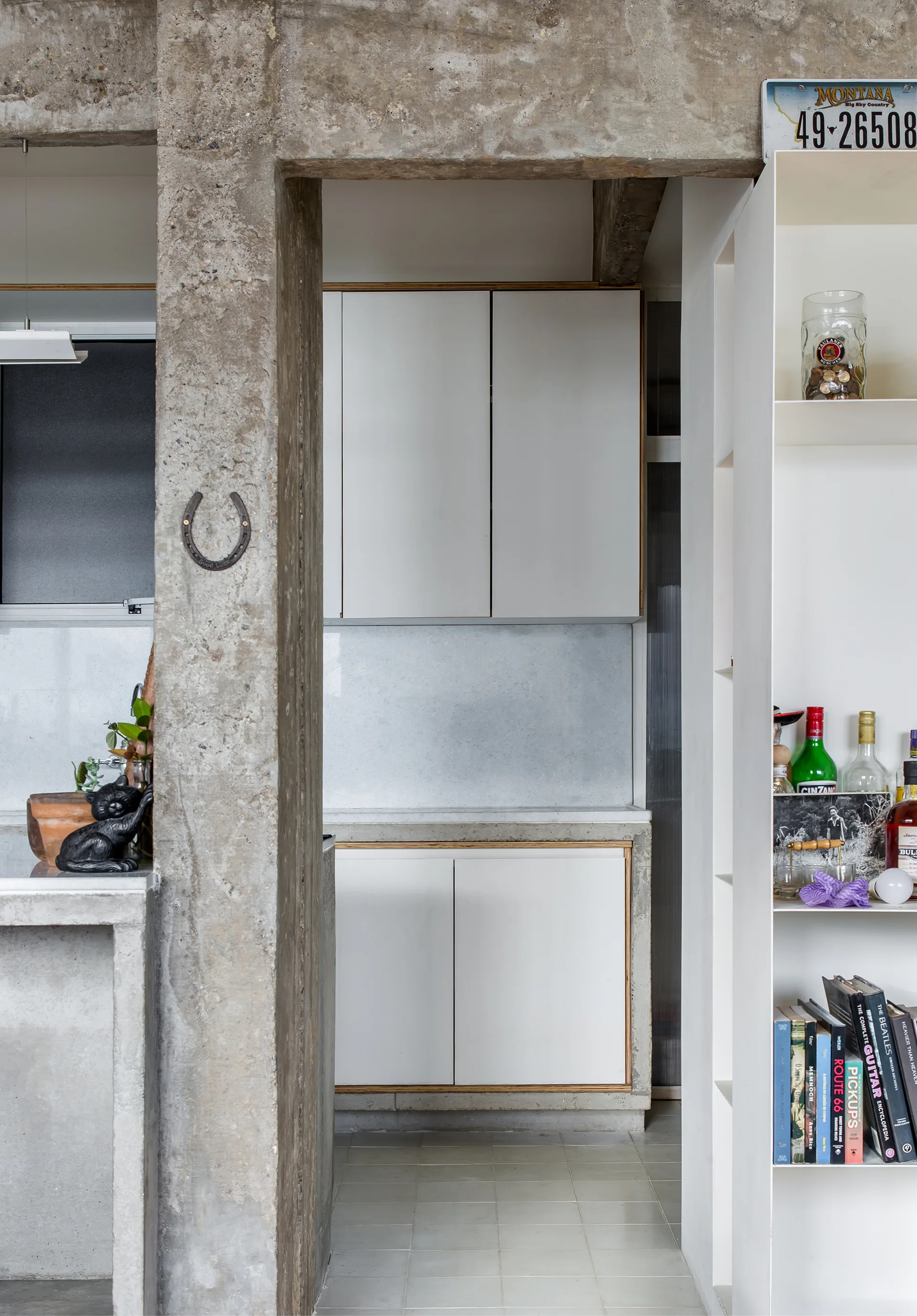 A raw concrete pillar frames a minimalist kitchen featuring flat-front white cabinets, light tile flooring, and a tall white open shelving unit.