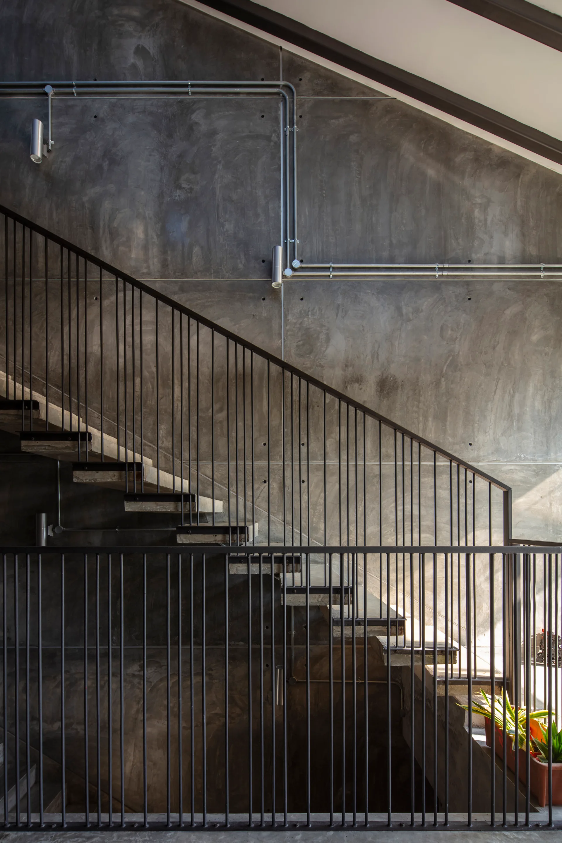 An industrial staircase with open concrete risers and vertical black metal railings rises against a smooth grey concrete wall with exposed metal conduit.