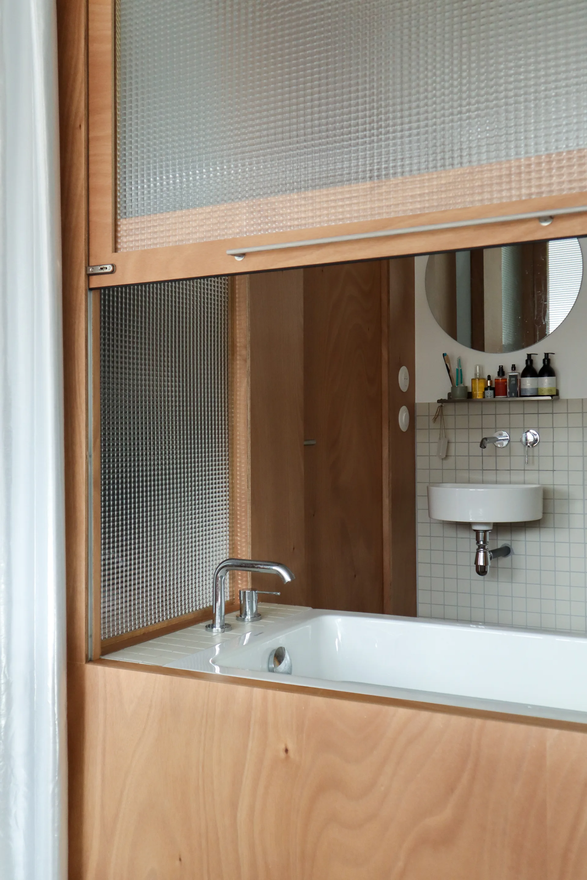A bathroom features a wooden partition with a bathtub, textured glass panel, and a wall-mounted sink against square white tiles.