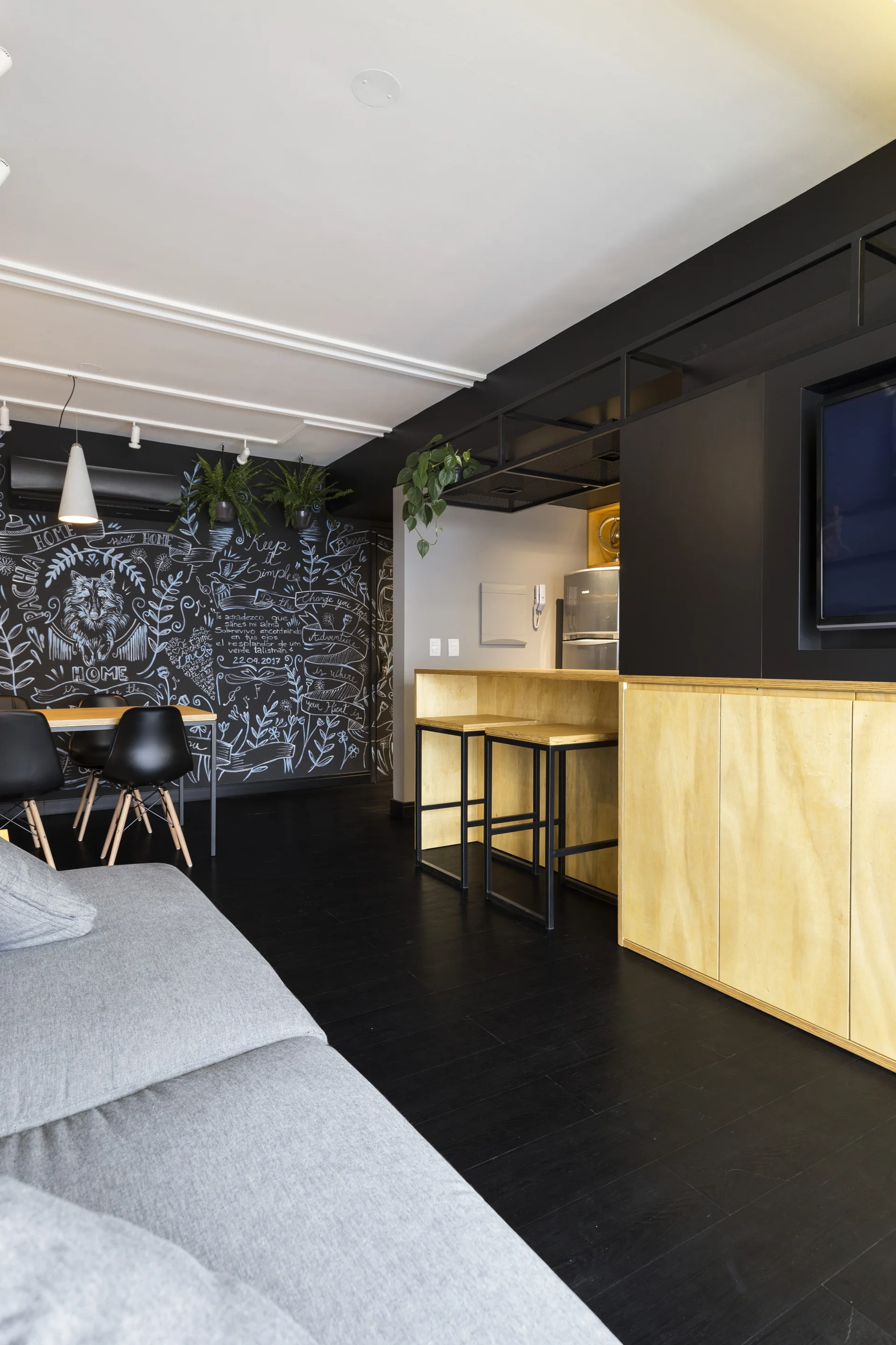 A modern open-plan living area features a black chalkboard wall with hand-drawn illustrations, light plywood kitchen cabinetry, and dark flooring.