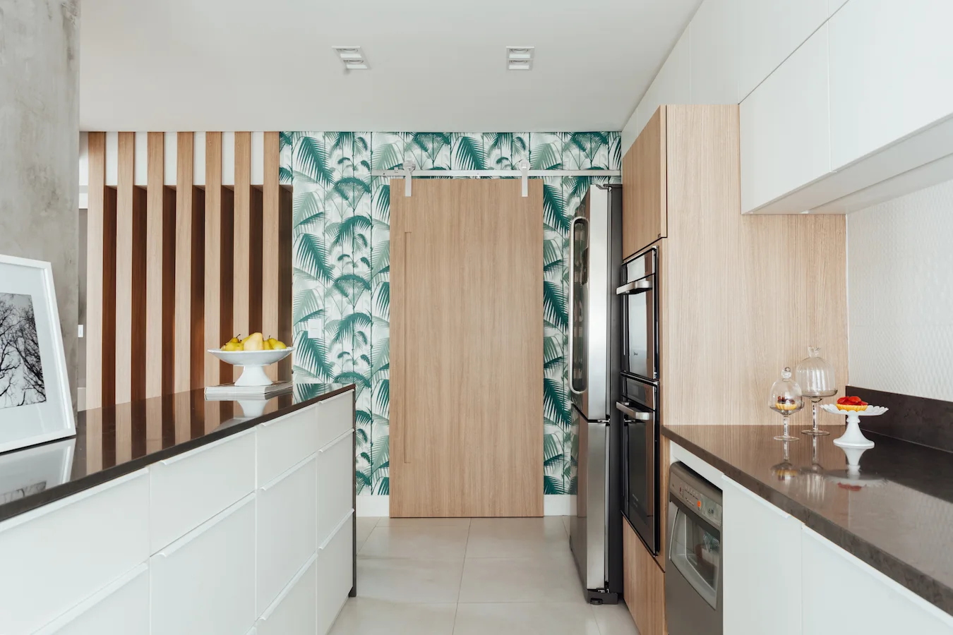 A modern kitchen featuring white handleless cabinetry, a wooden sliding barn door on a tropical palm-print wallpaper wall, and a wood slat divider.