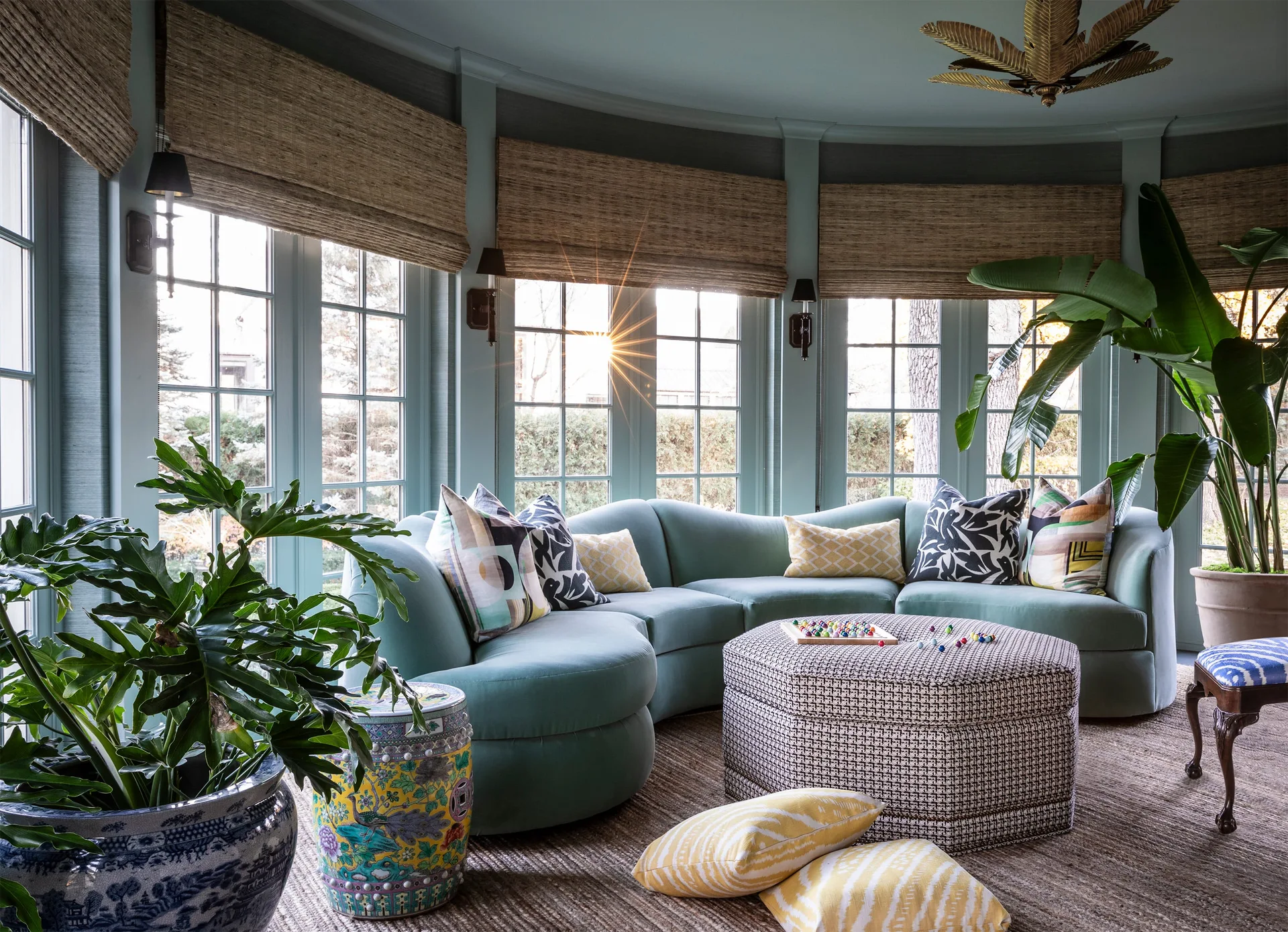 A curved teal sectional sofa sits in a sunroom with matching teal walls, large grid windows, woven roman shades, and a patterned hexagonal ottoman.