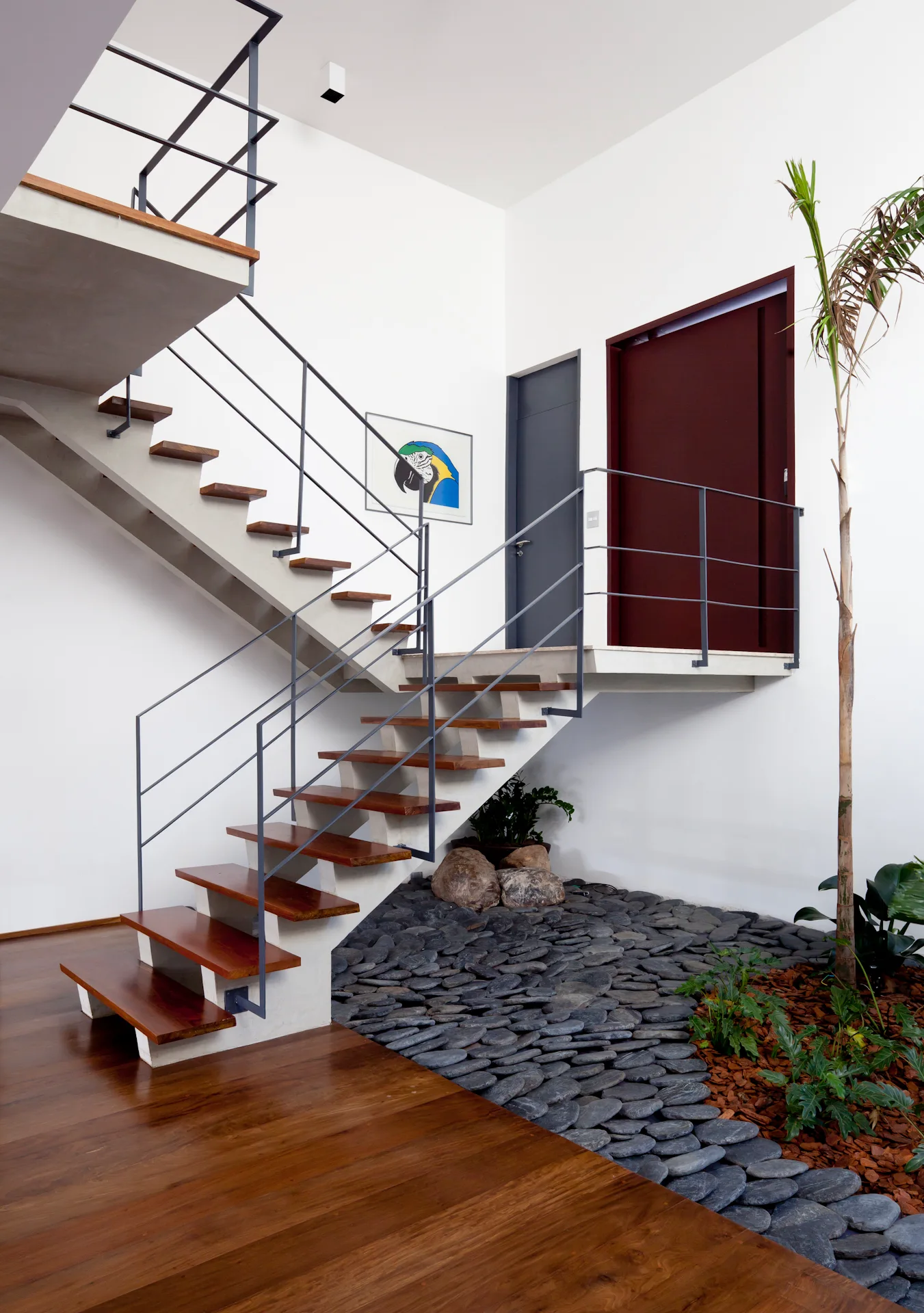 A modern floating staircase with wood treads and grey metal railings stands over an indoor garden bed of grey river stones.