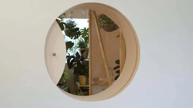 A circular wood-framed mirror with a partial sliding panel mounted on a white wall reflects lush potted plants in a bathroom.