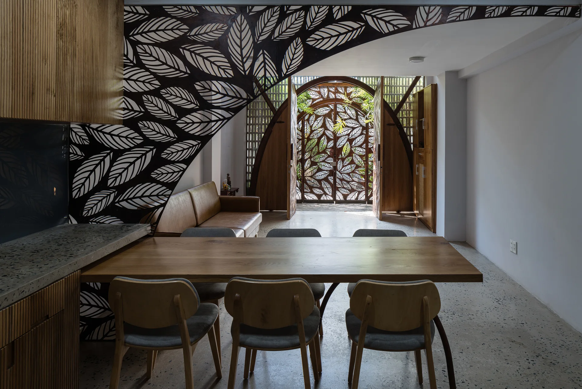 A long wooden dining table and chairs sit on terrazzo floors near a black leaf-patterned metal screen and an arched wooden entryway.