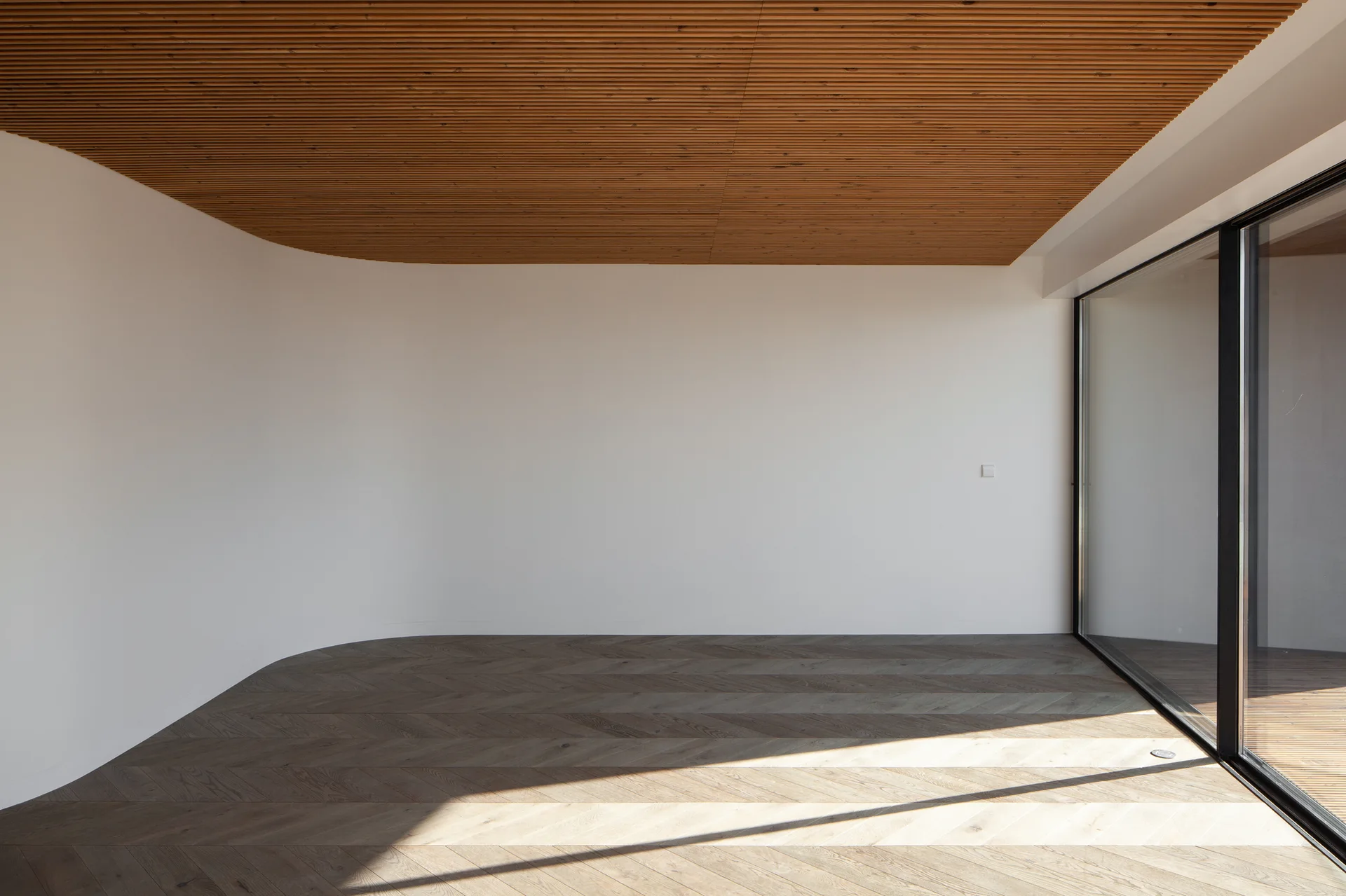 An empty room featuring a curved white wall, a slatted timber ceiling, herringbone patterned wood flooring, and a large sliding glass door.