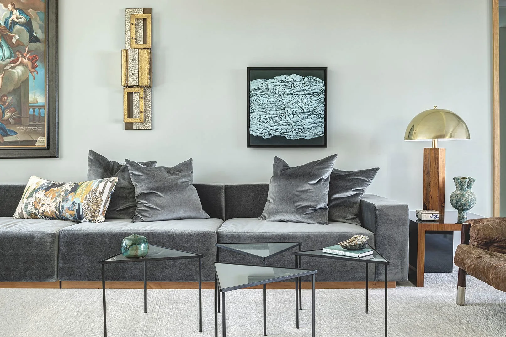 A gray velvet sofa is styled with matching throw pillows, triangular glass nesting tables, and a brass dome table lamp against a light gray wall.