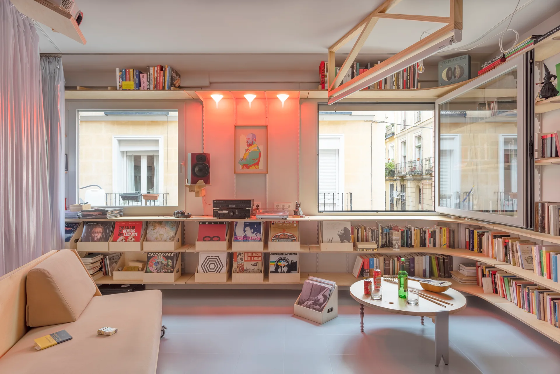 A living room with custom light wood shelving holding records and books under two windows, with a light-colored sofa and round coffee table.