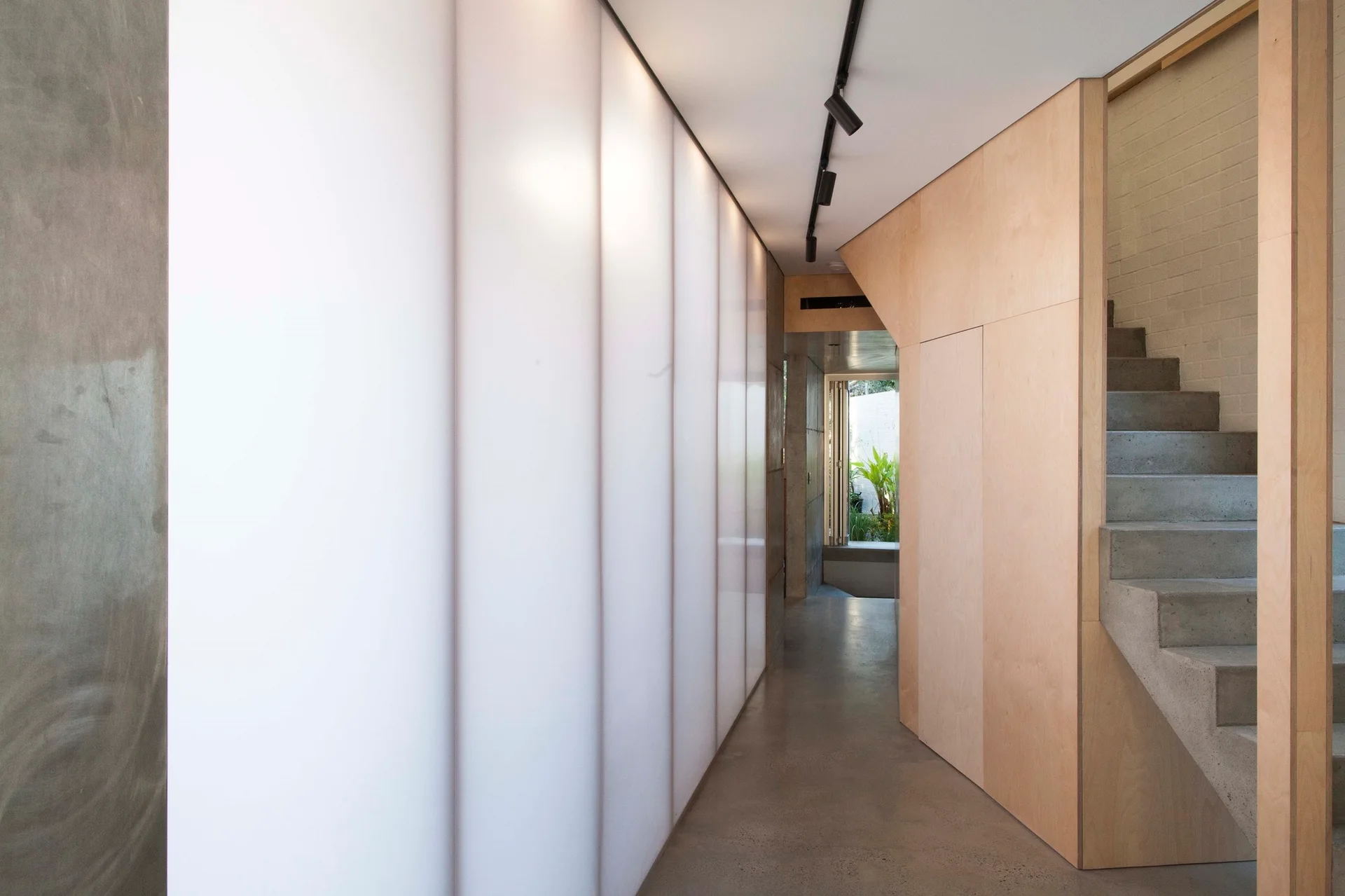 A modern hallway features a glowing backlit translucent wall panel, light wood storage cabinets, concrete floors, and concrete stairs.