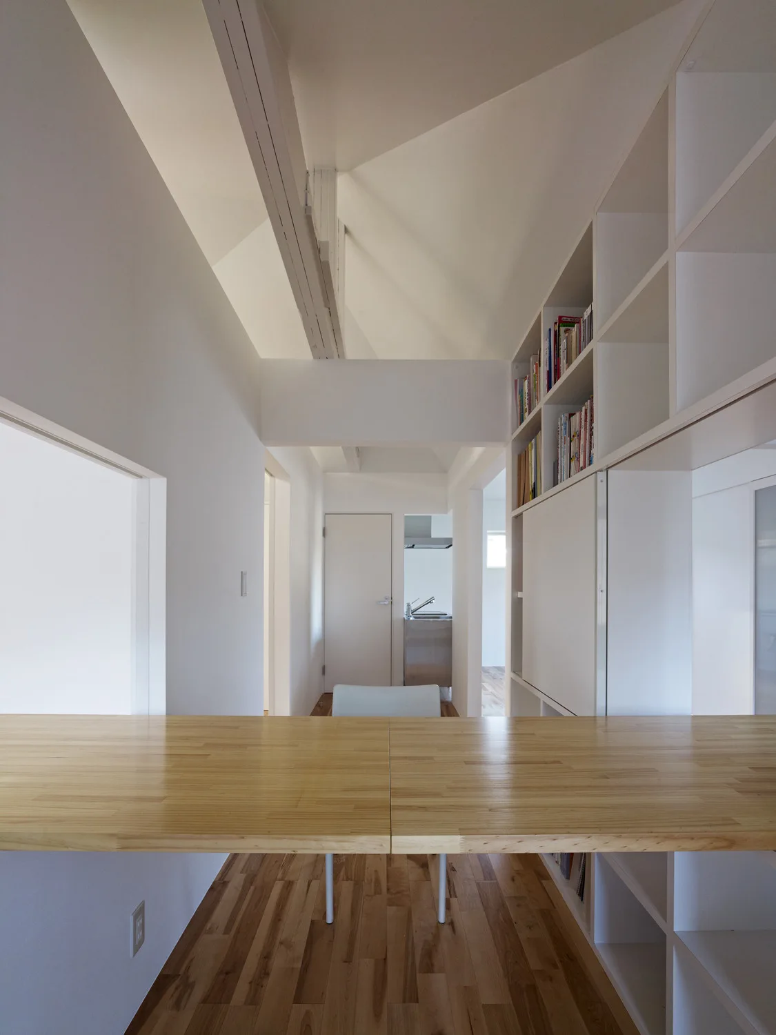 A minimalist workspace features a light wood desk counter in the foreground, white built-in shelving on the right, and wooden plank flooring.