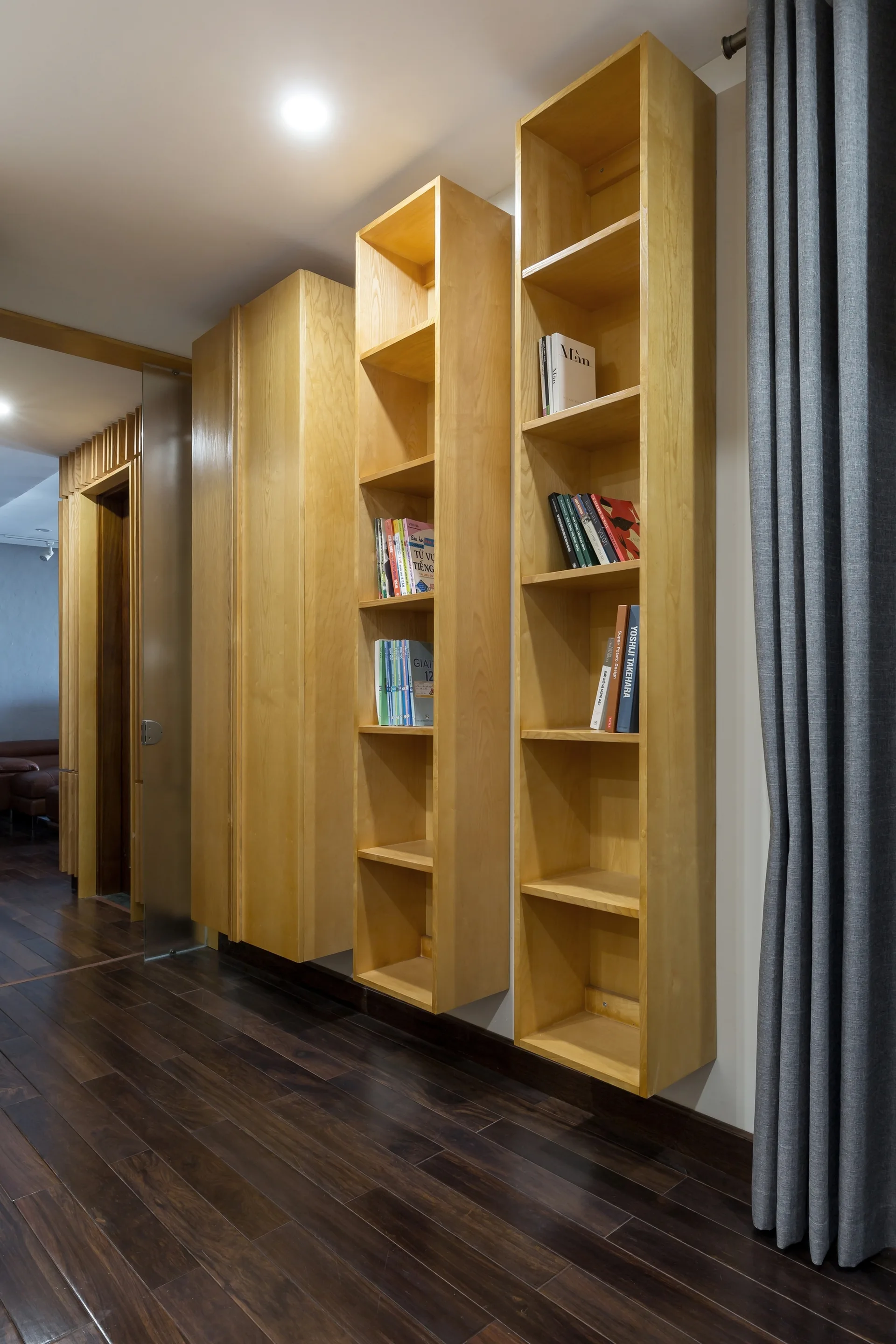 Tall wall-mounted light wood bookshelves hang in a hallway with dark wood flooring next to a grey curtain.