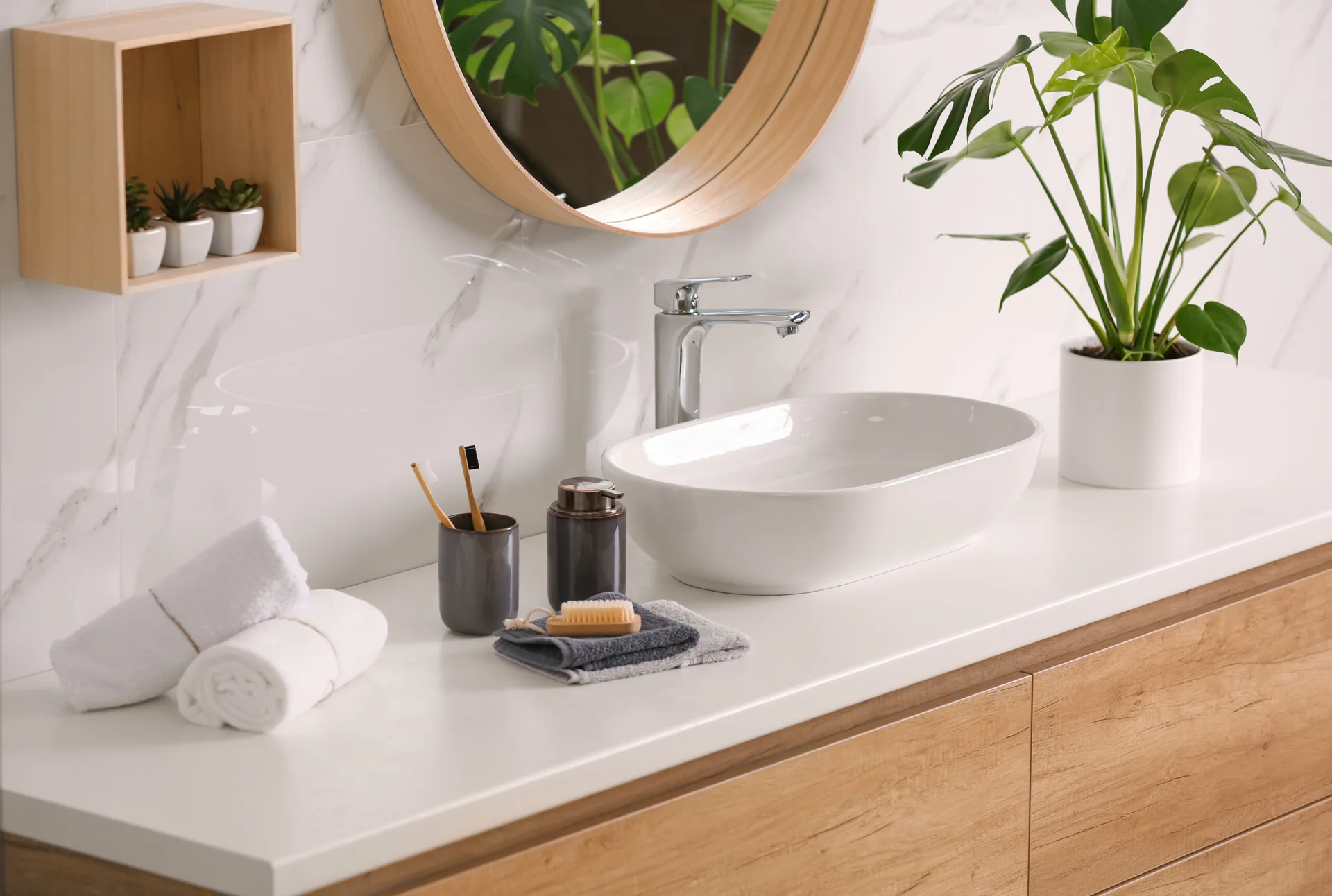 A modern bathroom vanity features a white vessel sink, light wood cabinetry, a round wood-framed mirror, and glossy marble wall tiles.