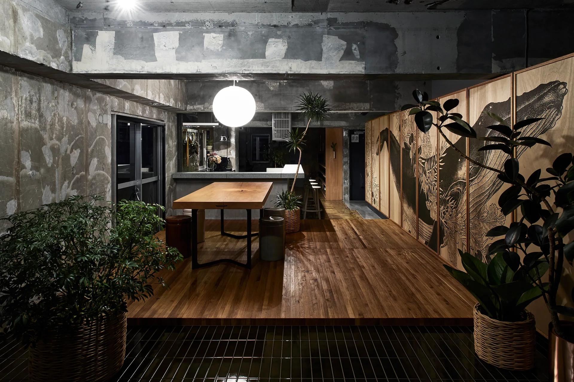 An industrial dining area with a wood table on a raised platform, surrounded by raw concrete walls and wood cabinet doors printed with a graphic mural.