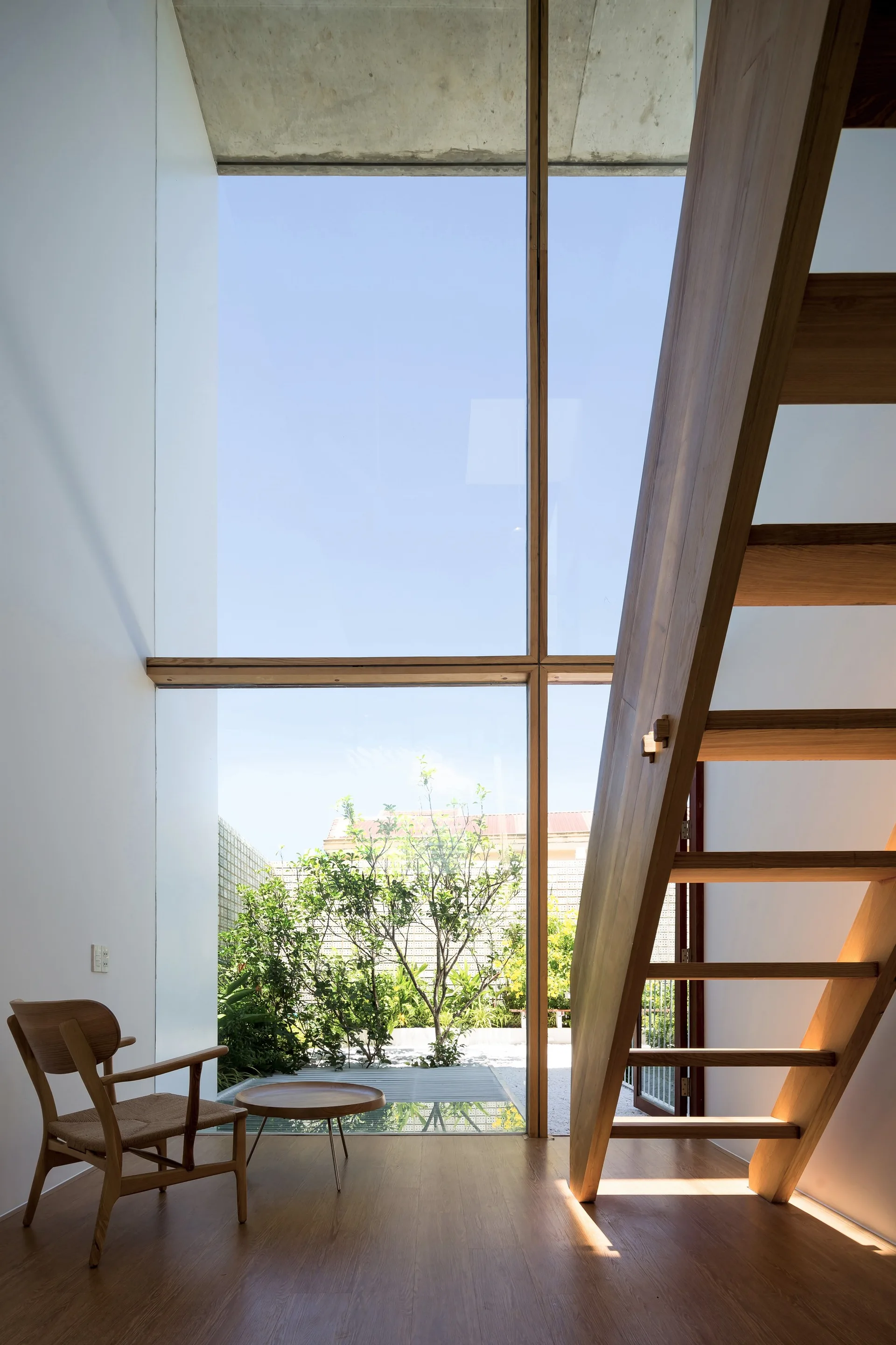 A minimalist room featuring an open-riser wooden staircase next to a towering glass wall overlooking a green courtyard.