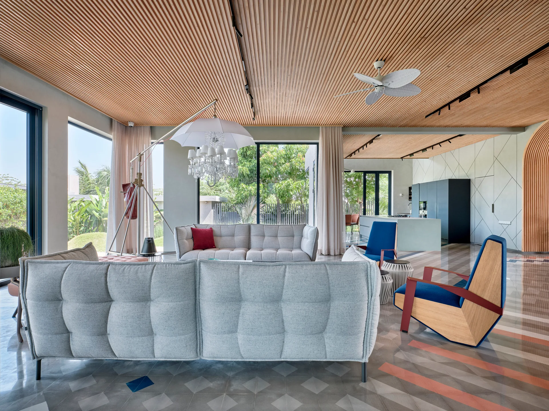 A modern living room features tufted grey sofas, blue armchairs, a patterned tile floor, and a linear wood slatted ceiling.