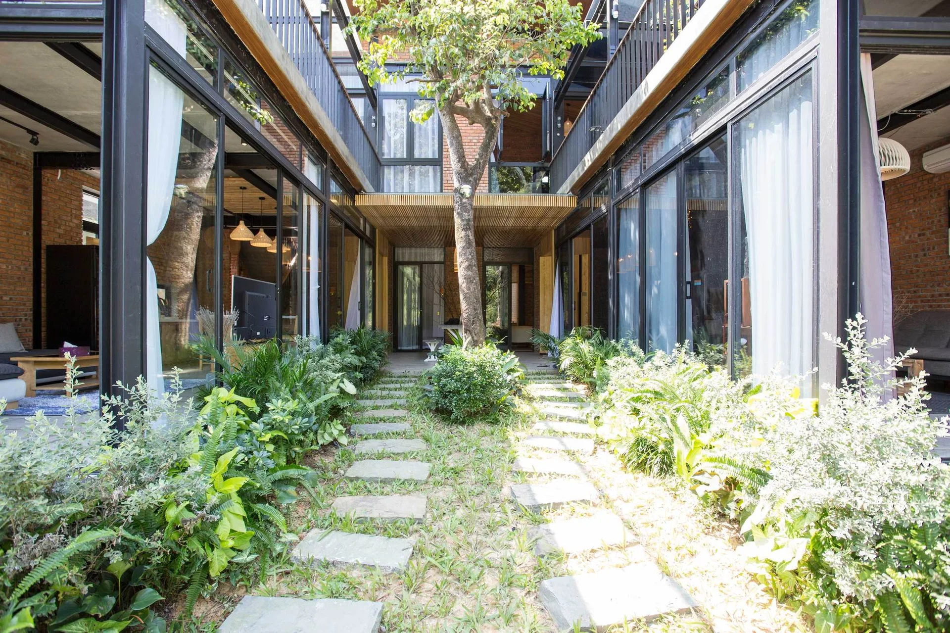 An outdoor residential courtyard features a stone stepping path, lush green plants, and a central tree surrounded by modern black metal glass walls.