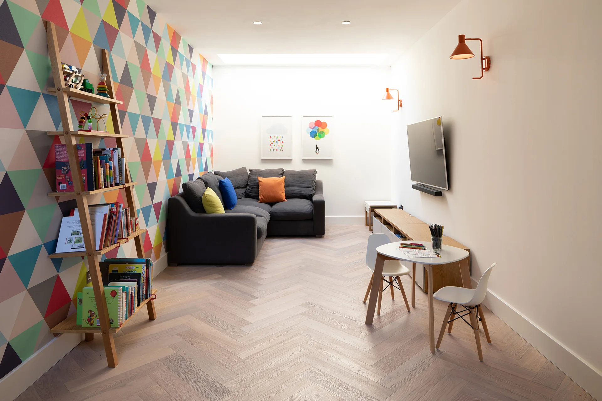 A family playroom with a colorful geometric triangle accent wall, a gray sectional sofa, herringbone wood floors, and a small white kids table.