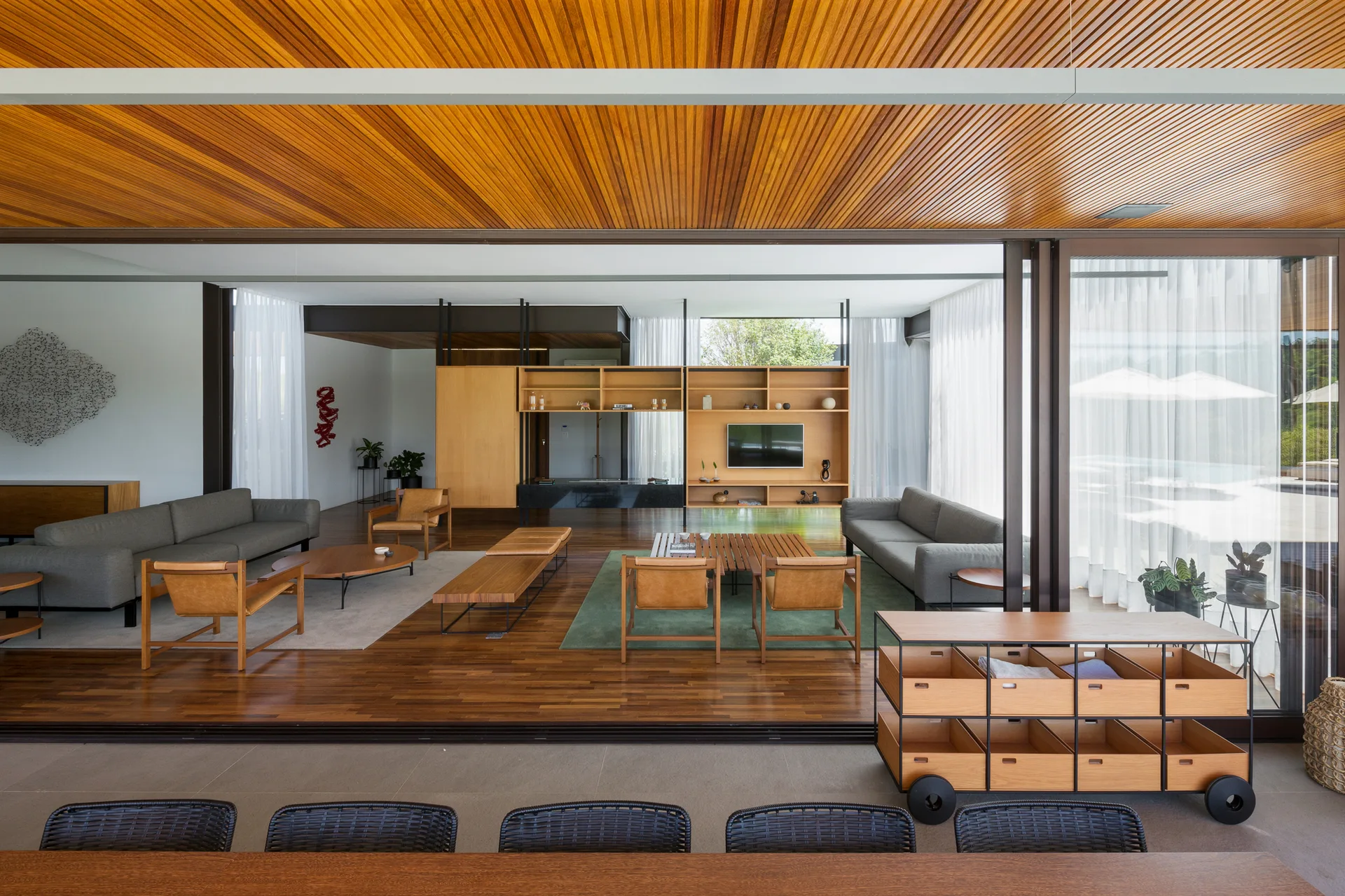 A spacious living room featuring a slatted wood ceiling, gray sofas, leather-accented armchairs, and a large central wood shelving unit partition.
