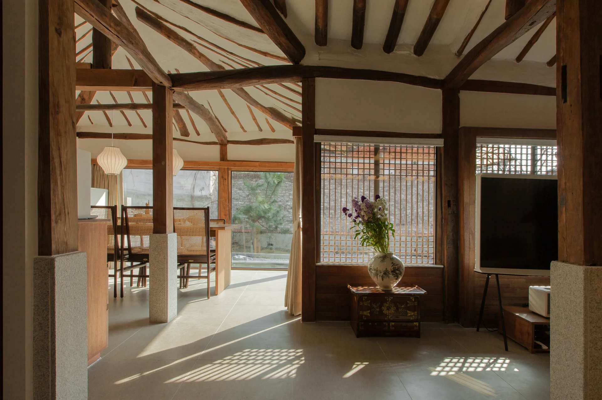 An interior room with exposed rustic wood rafters, stone-based columns, a lattice window screen, and a dining table with cane-back chairs.