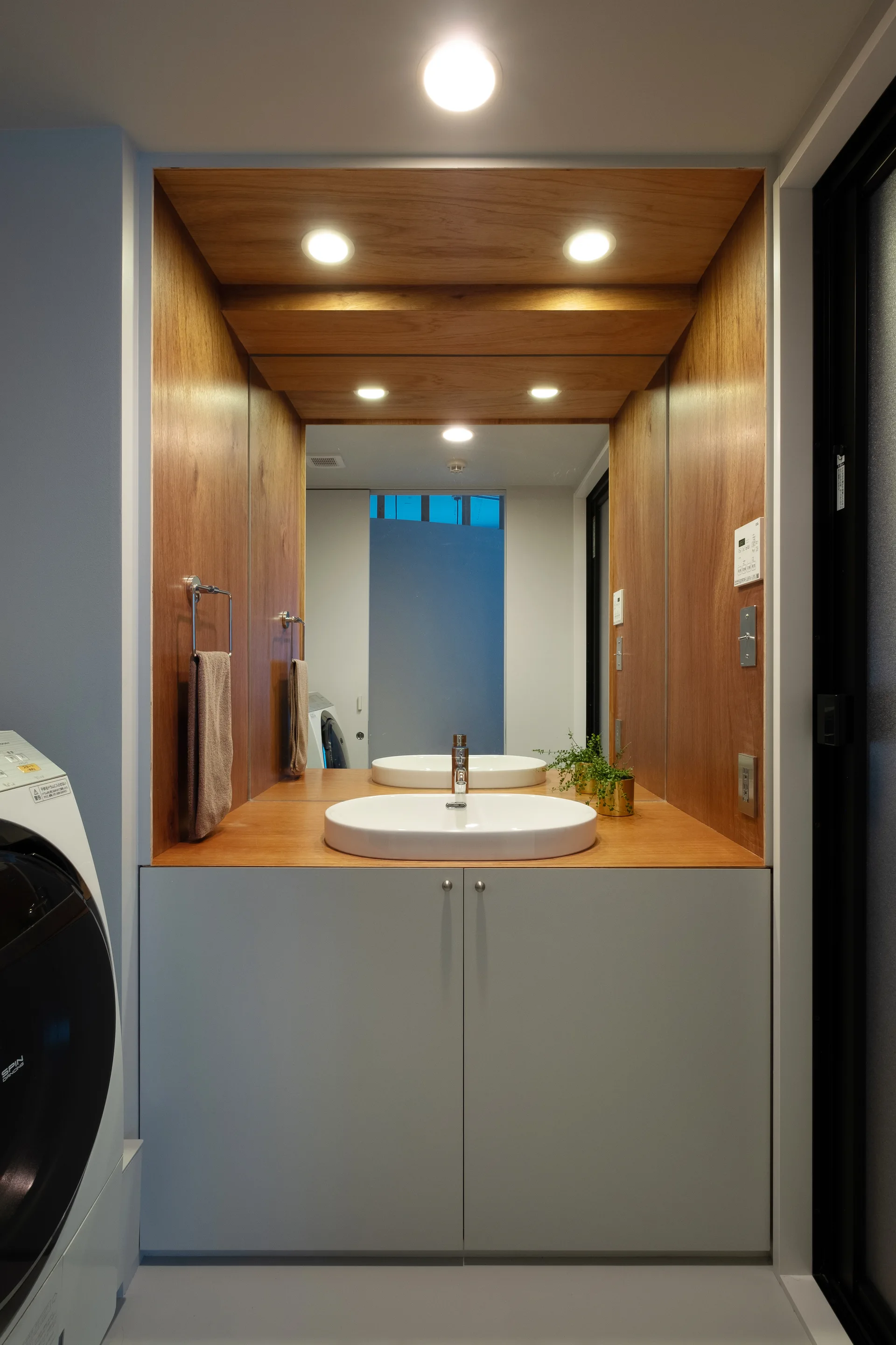 A bathroom vanity features a white vessel sink in a warm wood alcove with integrated lighting, a large mirror, and grey flat-panel cabinets.