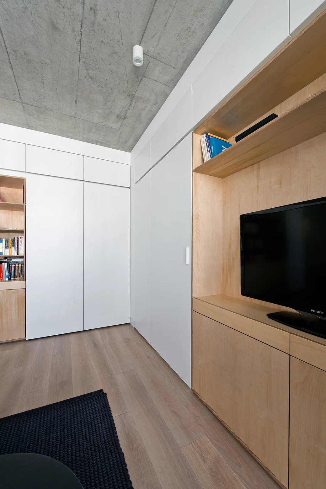 A modern living space features custom white and plywood built-in cabinetry, light wood floors, and a textured grey concrete ceiling.